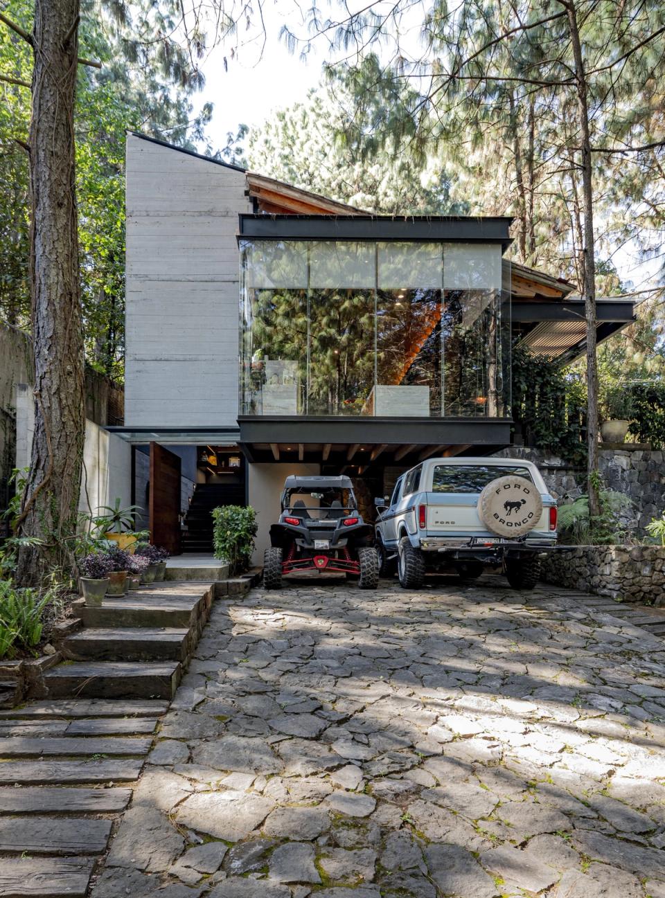 Fachada de concreto y cristal sobre la cochera, con camino de piedra entre pinos