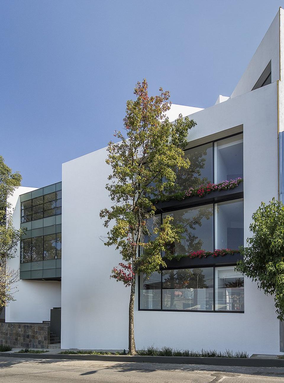 Fachada blanca con ventanales, jardineras con flores y un árbol al frente