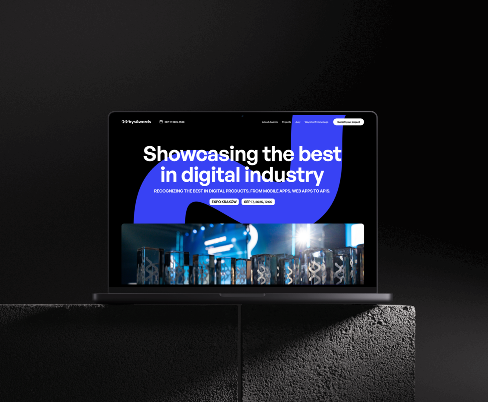 A laptop displays a modern website design featuring the text "Showcasing the best in digital industry" with a bold blue curved element and glass trophy imagery below.