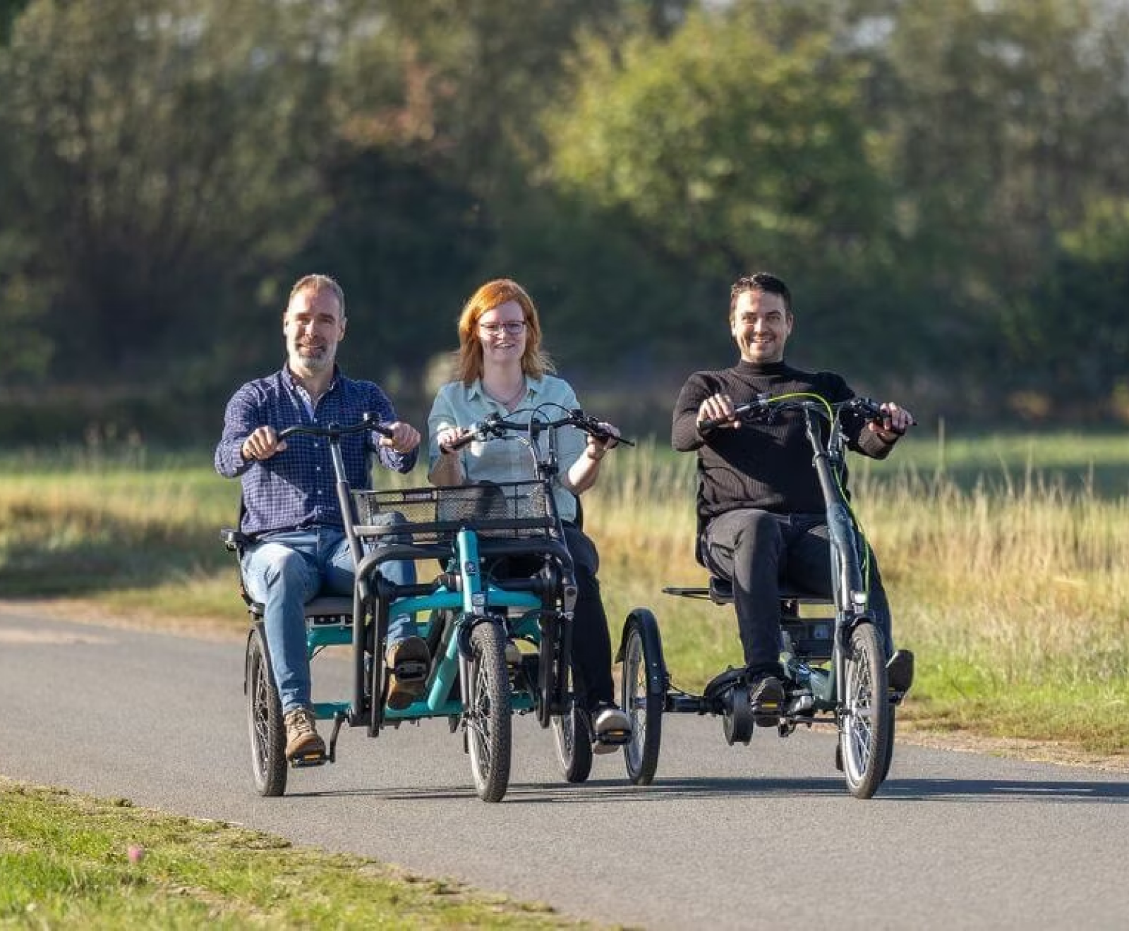 Three cyclists ride together on specialized adaptive tricycles along a sunny rural path.