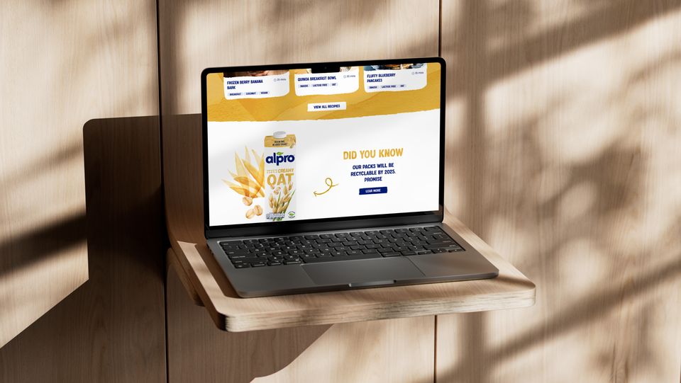 A laptop displaying an Alpro oat milk product webpage sits on a wooden floating shelf with dramatic shadow patterns cast on the wall.