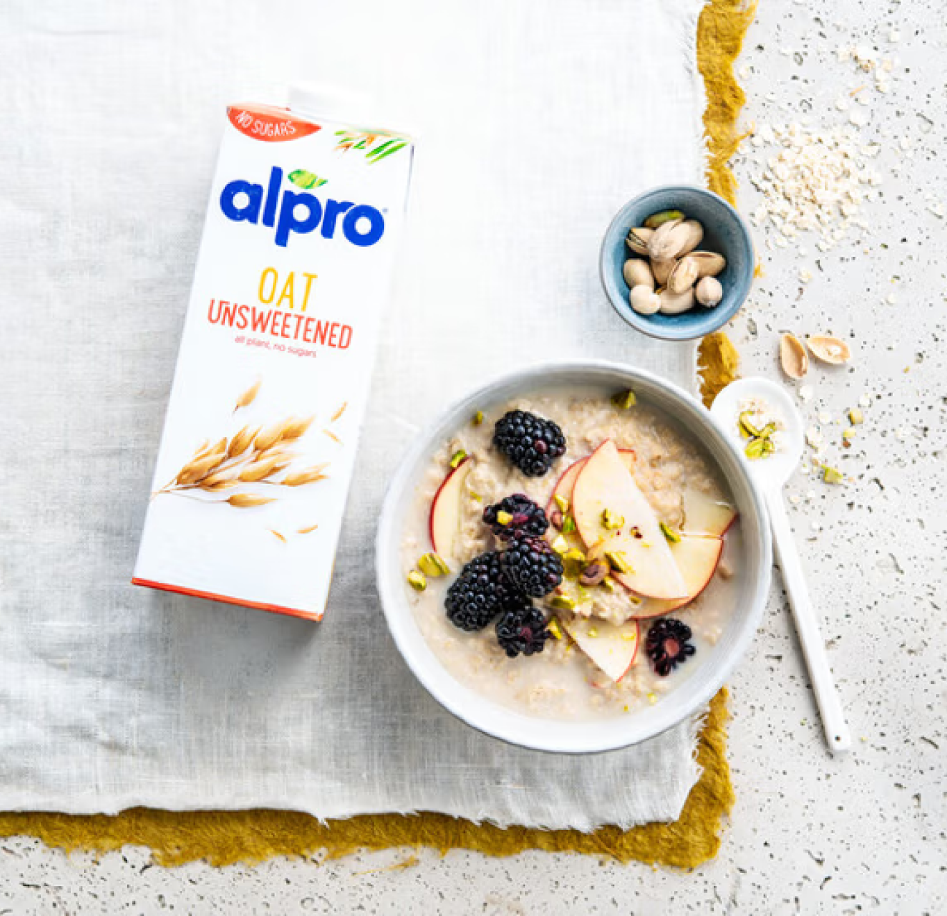 A bowl of oatmeal topped with fresh blackberries and apple slices sits alongside an Alpro unsweetened oat milk carton and a small dish of pistachios.