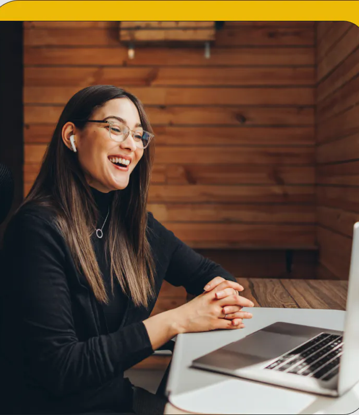 A cheerful professional wearing glasses and a black turtleneck sits at a wooden desk with a laptop in a warm, wood-paneled room.