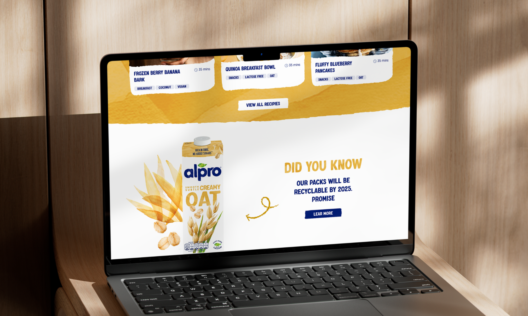 A laptop displays an Alpro oat milk product webpage featuring their sustainability promise about recyclable packaging by 2025.