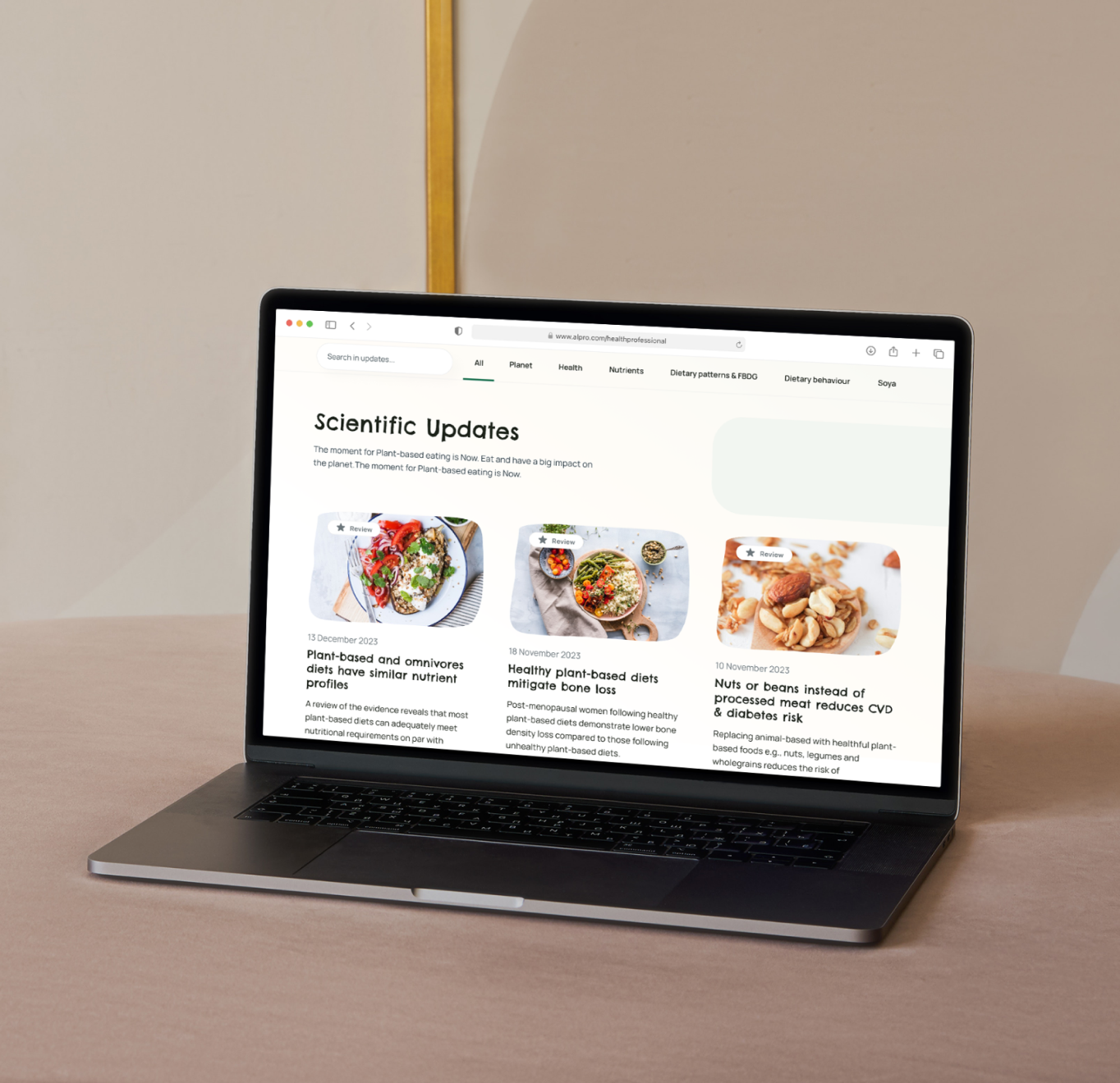 A MacBook displays a website's "Scientific Updates" section featuring three articles about plant-based diets, nutrition, and health benefits.