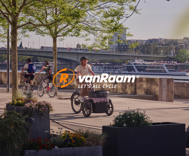 A cyclist rides a Van Raam special bike along a riverside path decorated with flower planters, with a bridge and boats visible in the background.