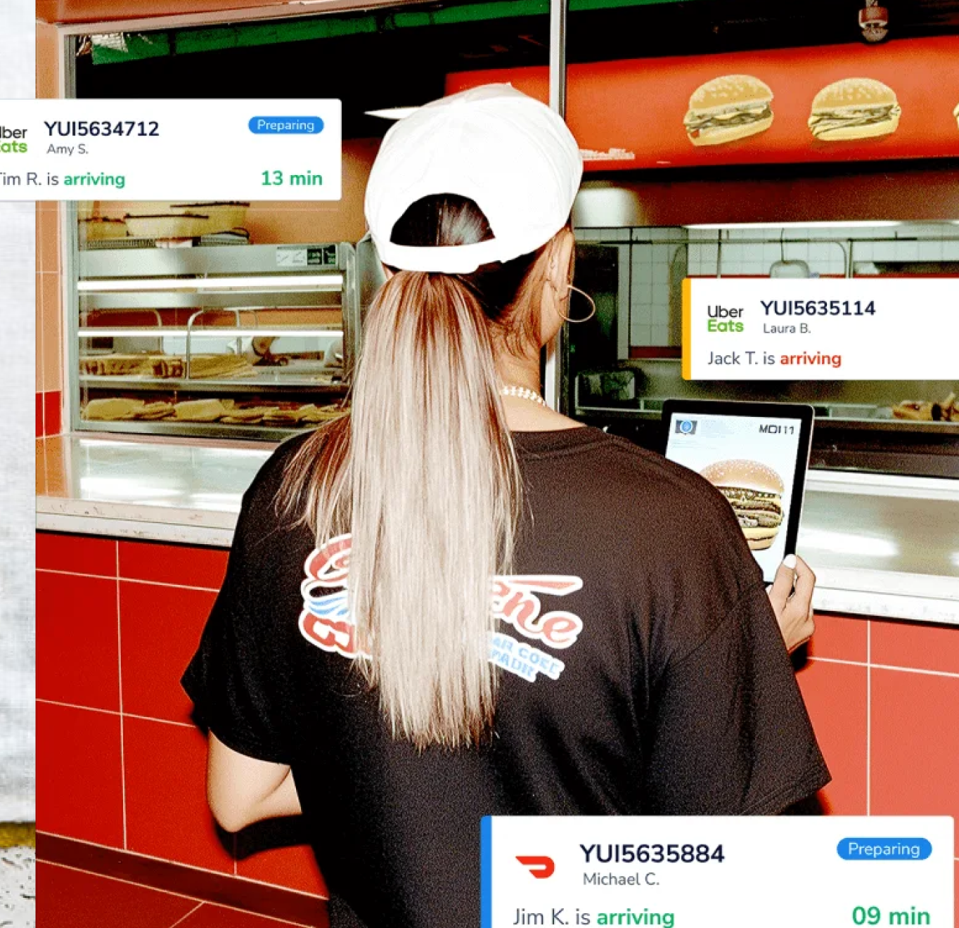 A restaurant worker with long blonde hair and a white cap checks multiple Uber Eats delivery orders on a screen at a burger counter.