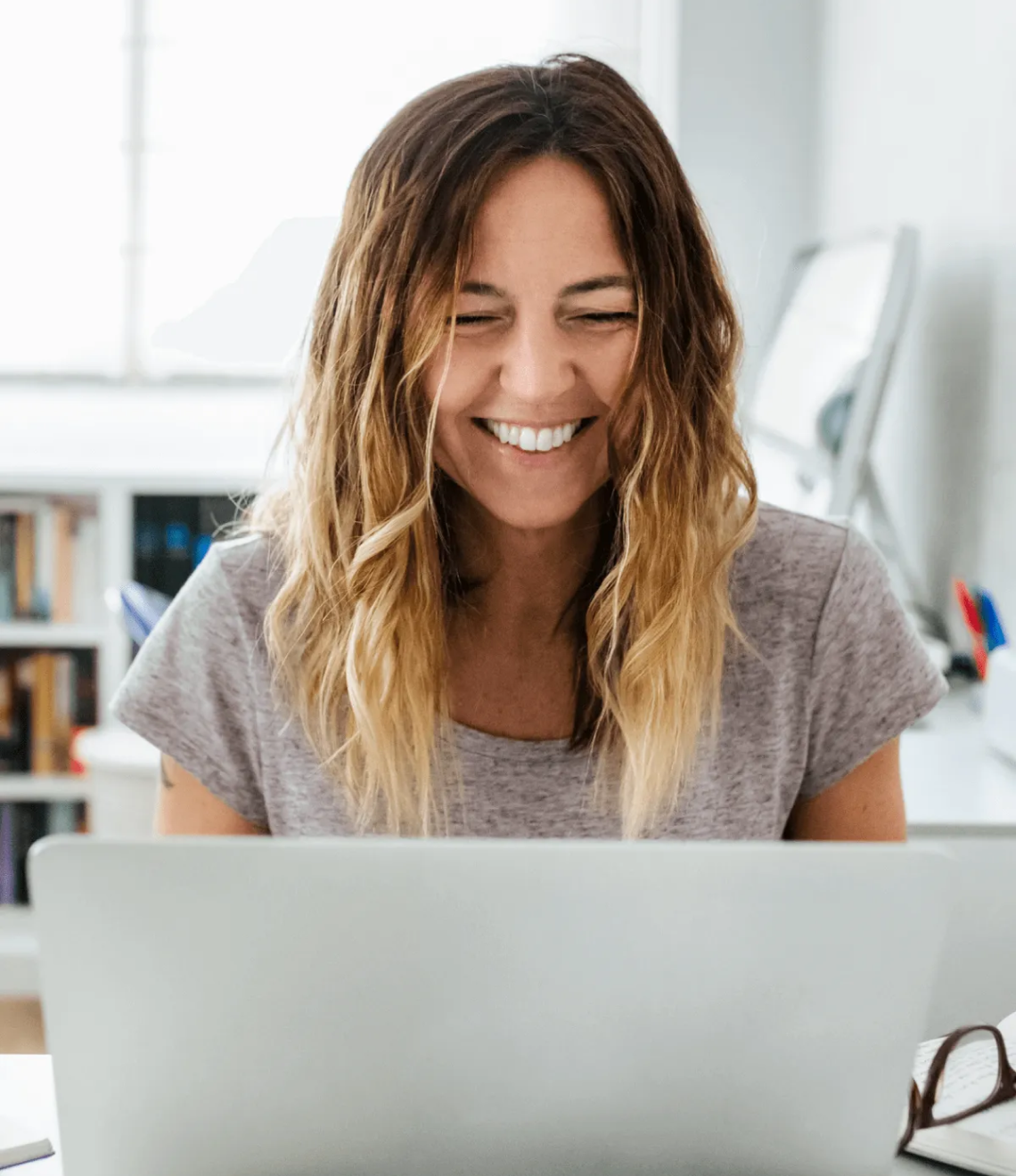 A person with ombre-styled hair and a grey t-shirt smiles warmly while working on a laptop in a bright office space.