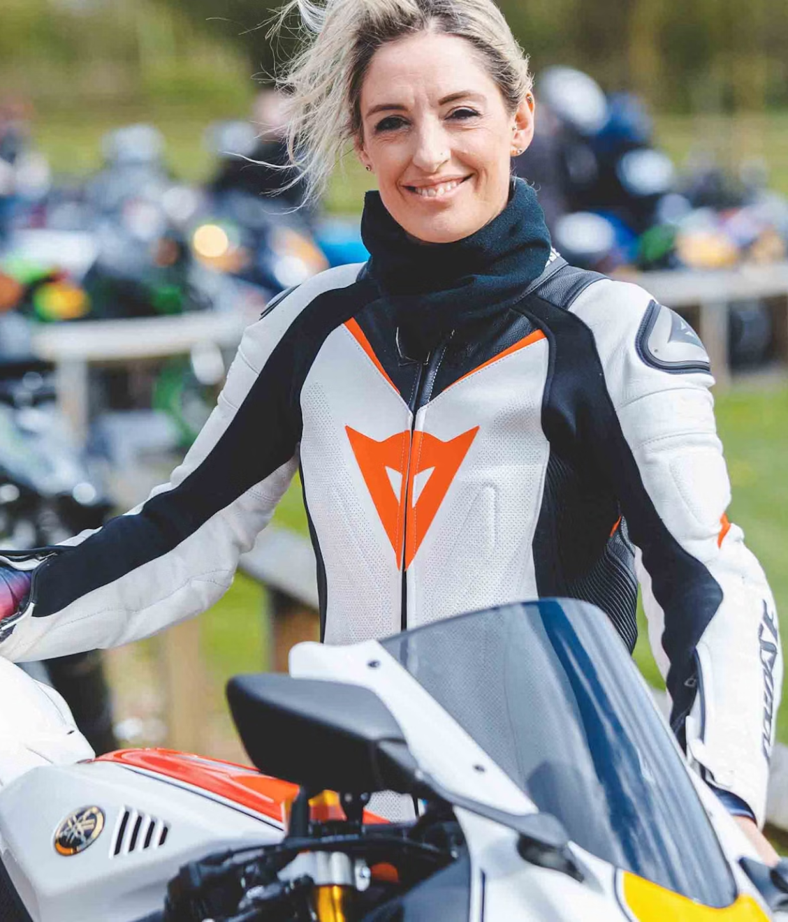 A motorcyclist wearing a white and orange racing suit stands beside their sport bike at what appears to be a racing or motorcycle event.