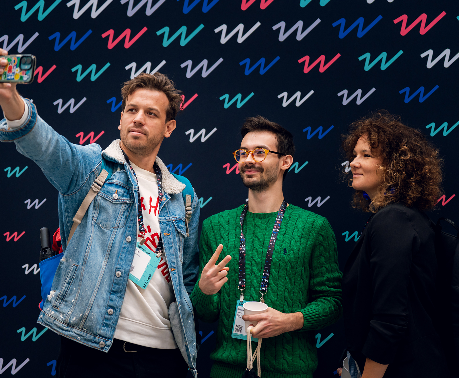 Three friends pose for a selfie against a patterned wall featuring colorful "W" letters, with one wearing a denim jacket, another in a green sweater, and the third in black.