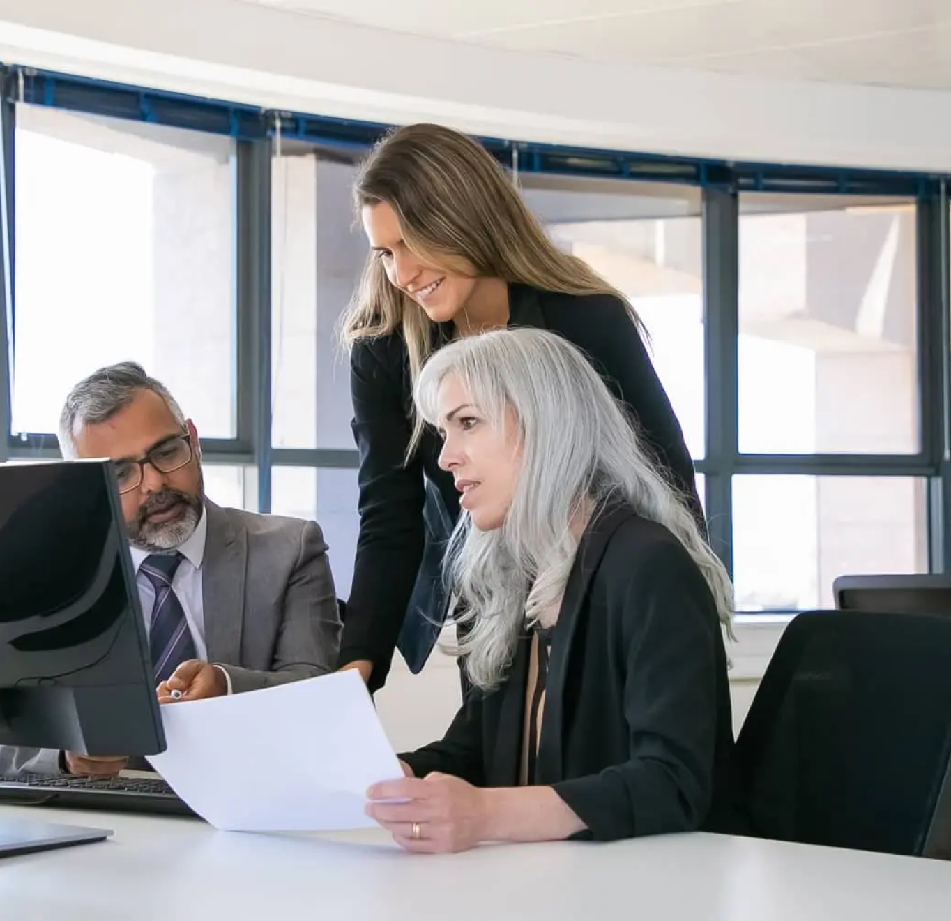 Three business professionals collaborate while reviewing documents at a modern office desk with large windows in the background.