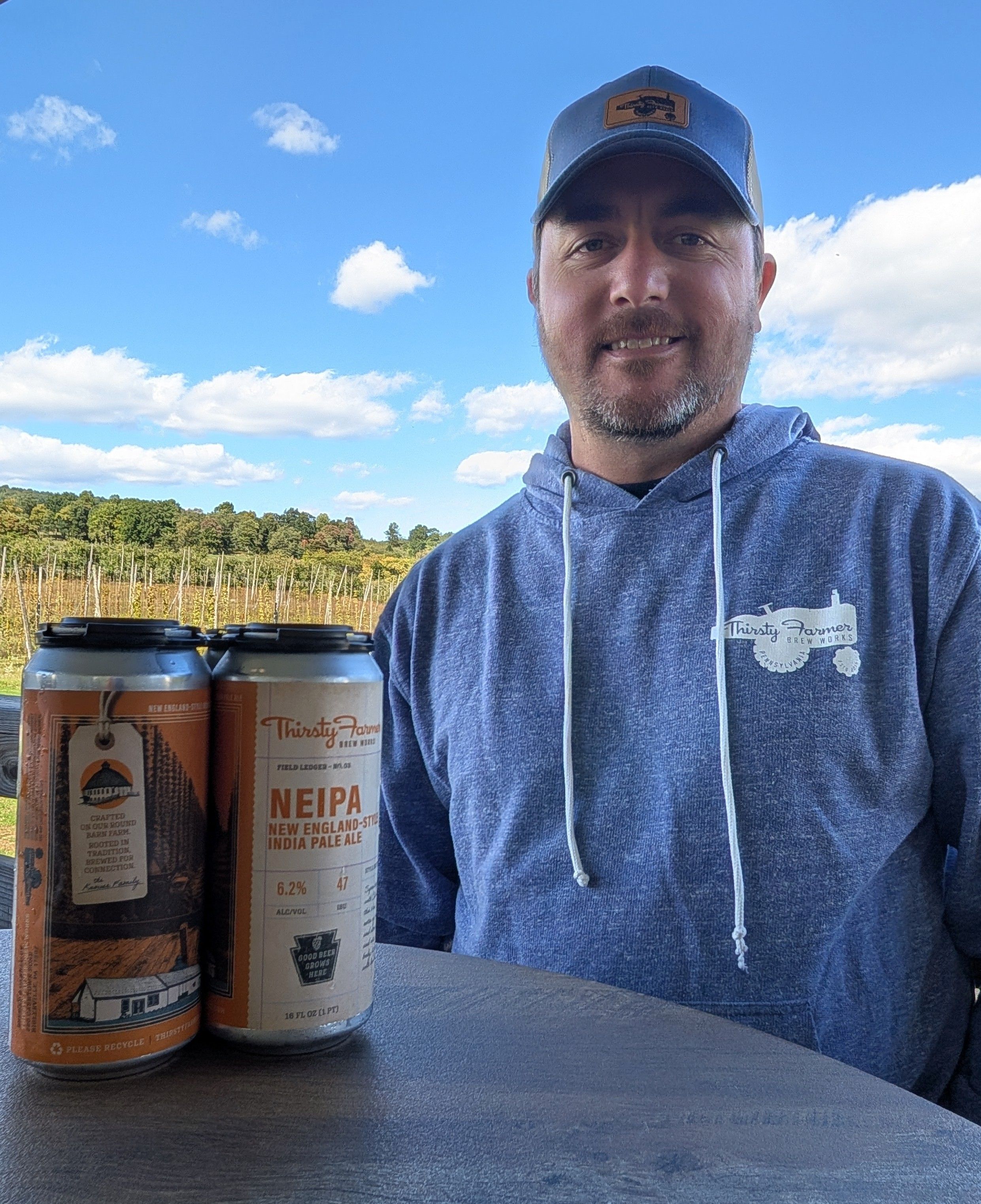 Kevin Knouse at Thirsty Farmer with two NEIPA cans, the hop yard behind him