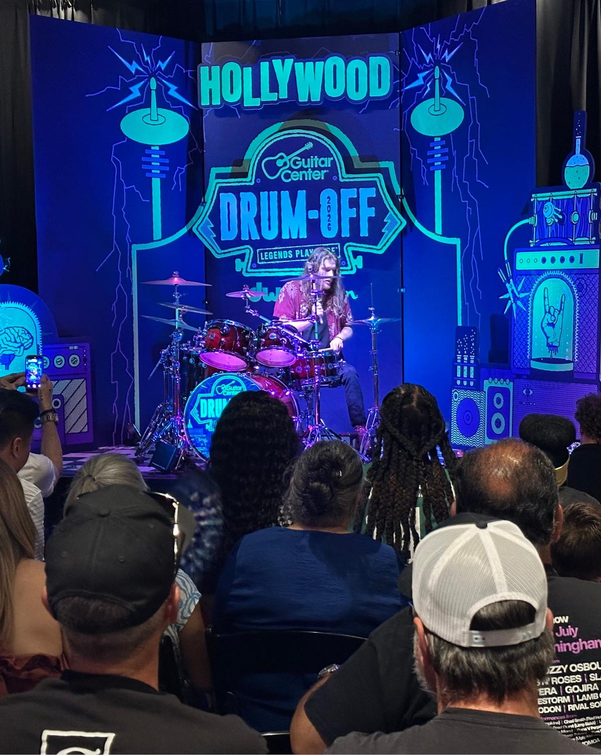 The Hollywood regional stage: lab machinery set pieces and lightning around a HOLLYWOOD Drum-Off sign, with a drummer playing to a crowd