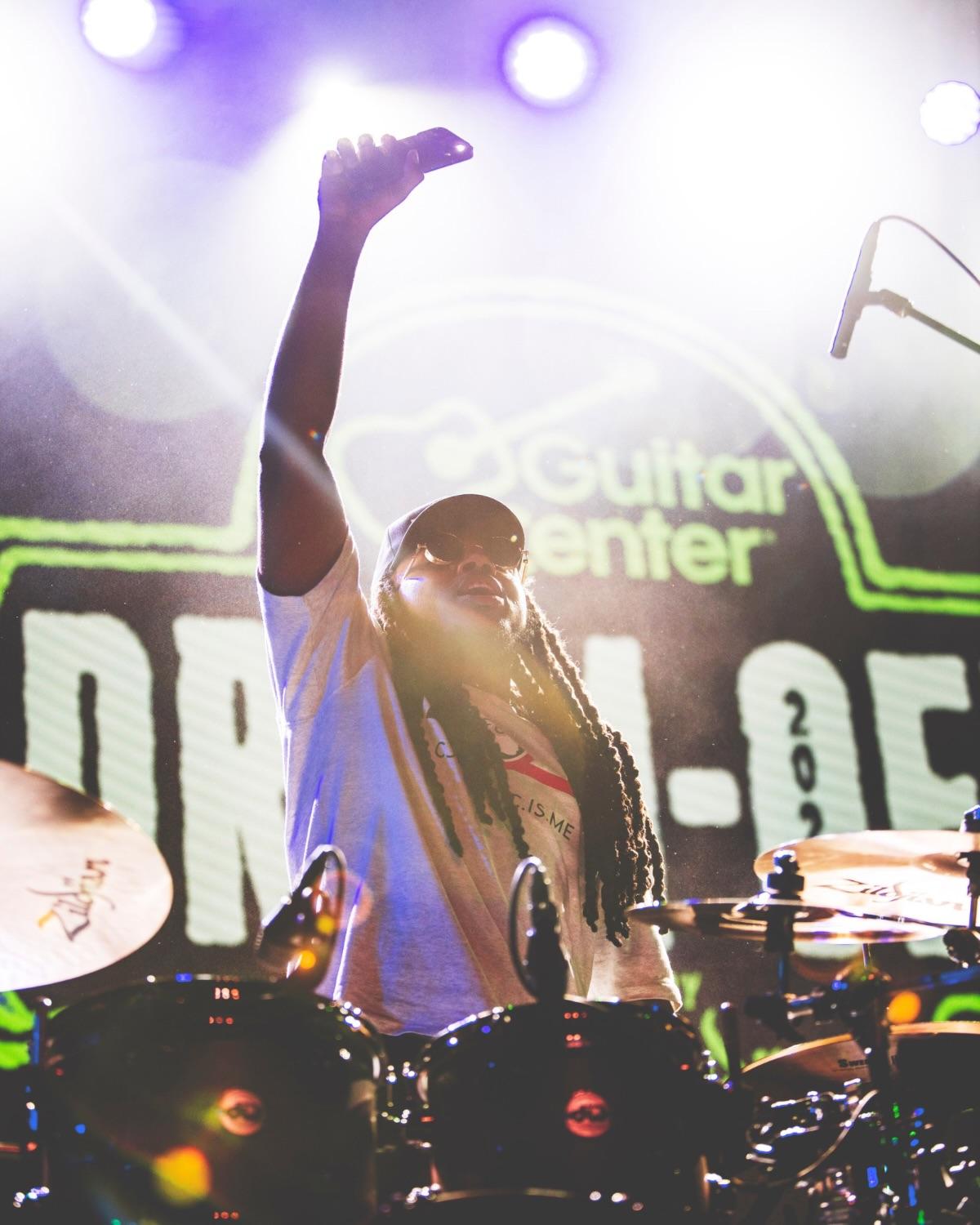A performer with a raised fist in front of a giant Drum-Off logo on the stage screen