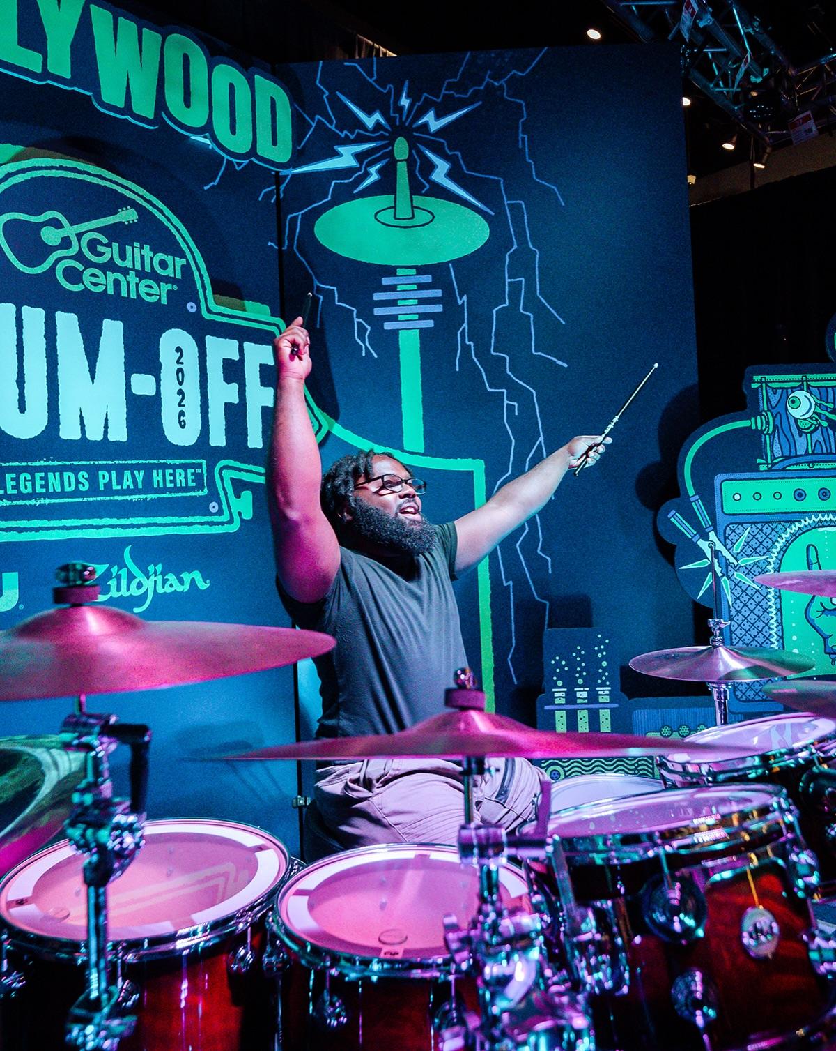 A drummer on the Hollywood stage surrounded by lab-equipment set pieces under a HOLLYWOOD Drum-Off sign