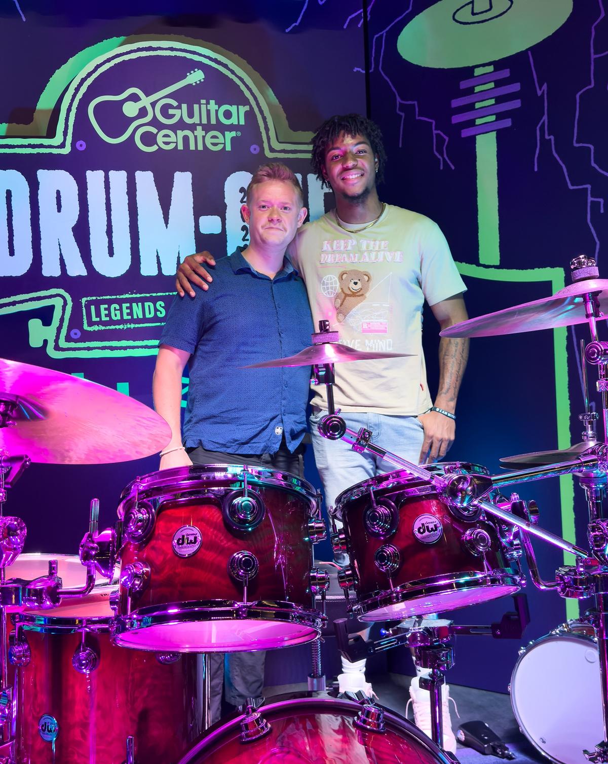 Two people posing on a regional Drum-Off stage set built from the lab art, under a Guitar Center Drum-Off sign