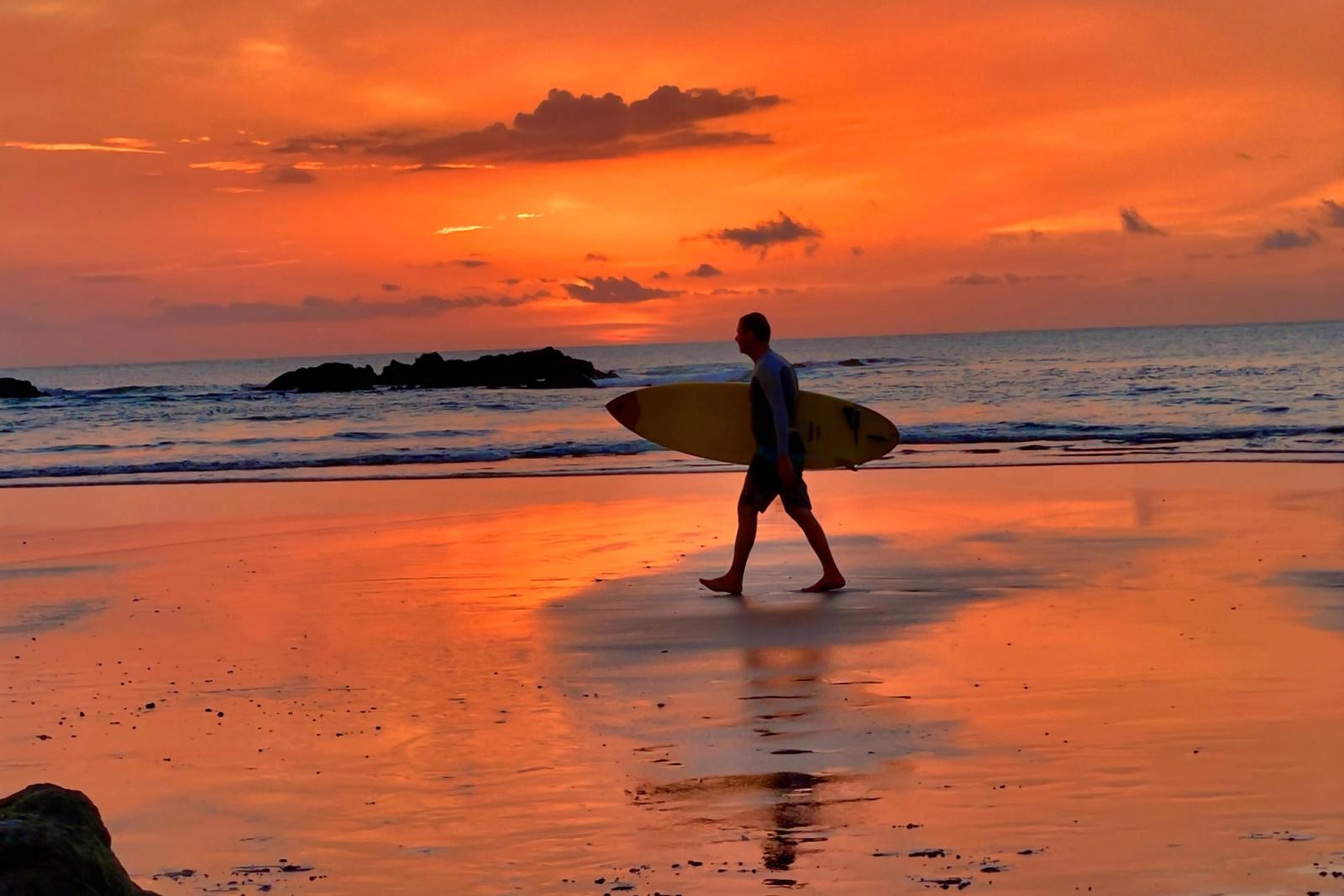 A surfer carrying a board along the beach at sunset in Tamarindo, Costa Rica