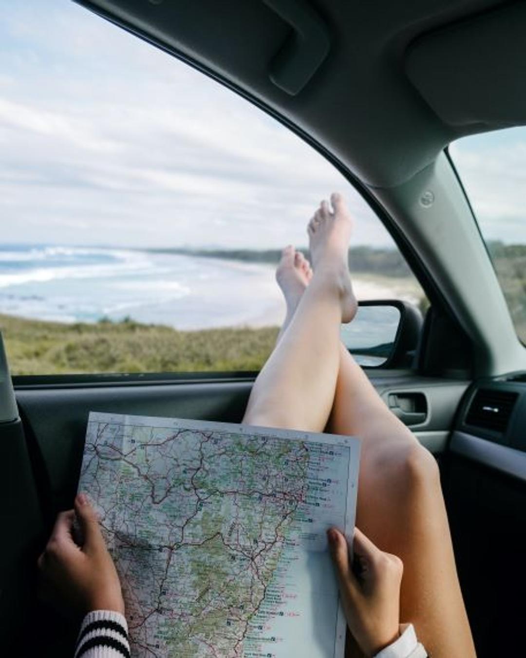 A car passenger is checking out a hand-held map for the best travel route.  The passenger is very relaxed as the passenger's feet are crossed and atop the passenger window ledge.