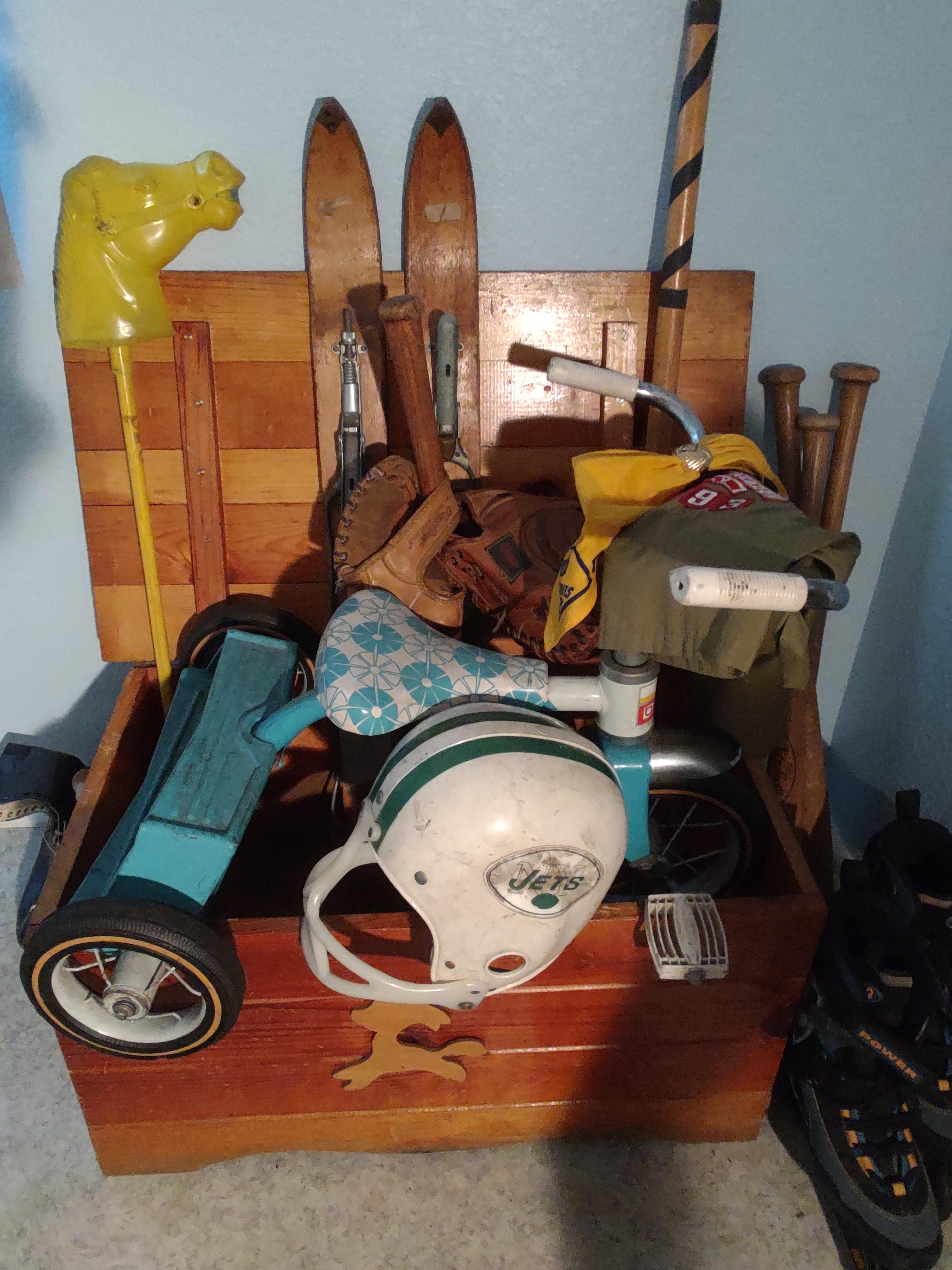 A picture of an old wooden varnished child's crate, containing many childhood sports toys and items, including an old pair of short wooden skis, a stickball bat, four wooden classic baseball bats, a couple of baseball gloves, a NY Jets plastic football helmet, a child's blue and white tricycle, and a pair of in-line skates from yesteryear.