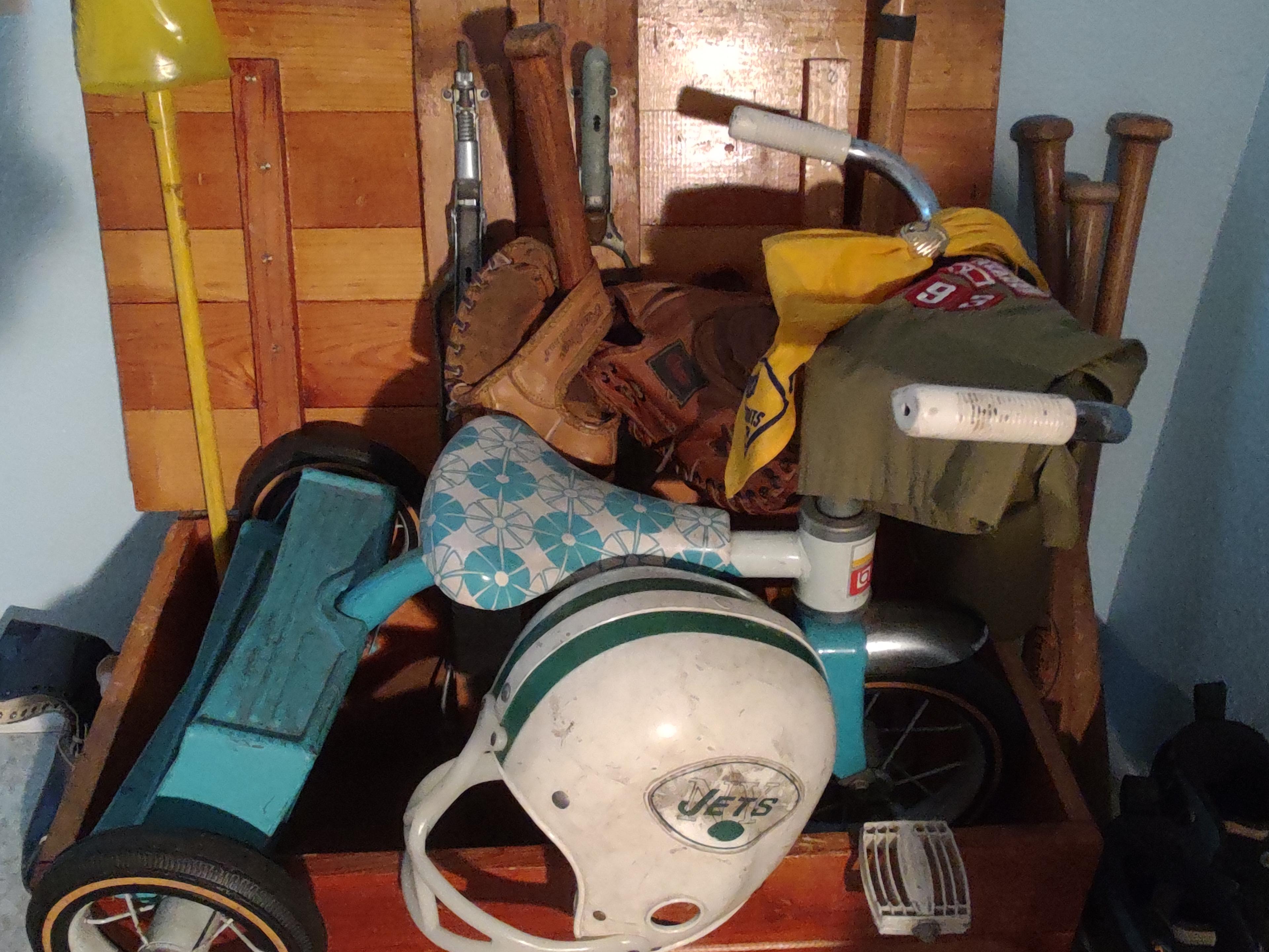 A picture of an old wooden varnished child's crate, containing many childhood sports toys and items, including an old pair of short wooden skis, a stickball bat, four wooden classic baseball bats, a couple of baseball gloves, a NY Jets plastic football helmet, a child's blue and white tricycle, and a pair of in-line skates from yesteryear.