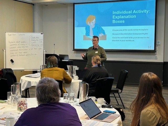 workshop in progress. a presenter stands below a projection headed Individual Activity: Explanation boxes. There is a full whiteboard next to them. A number of participants are visible