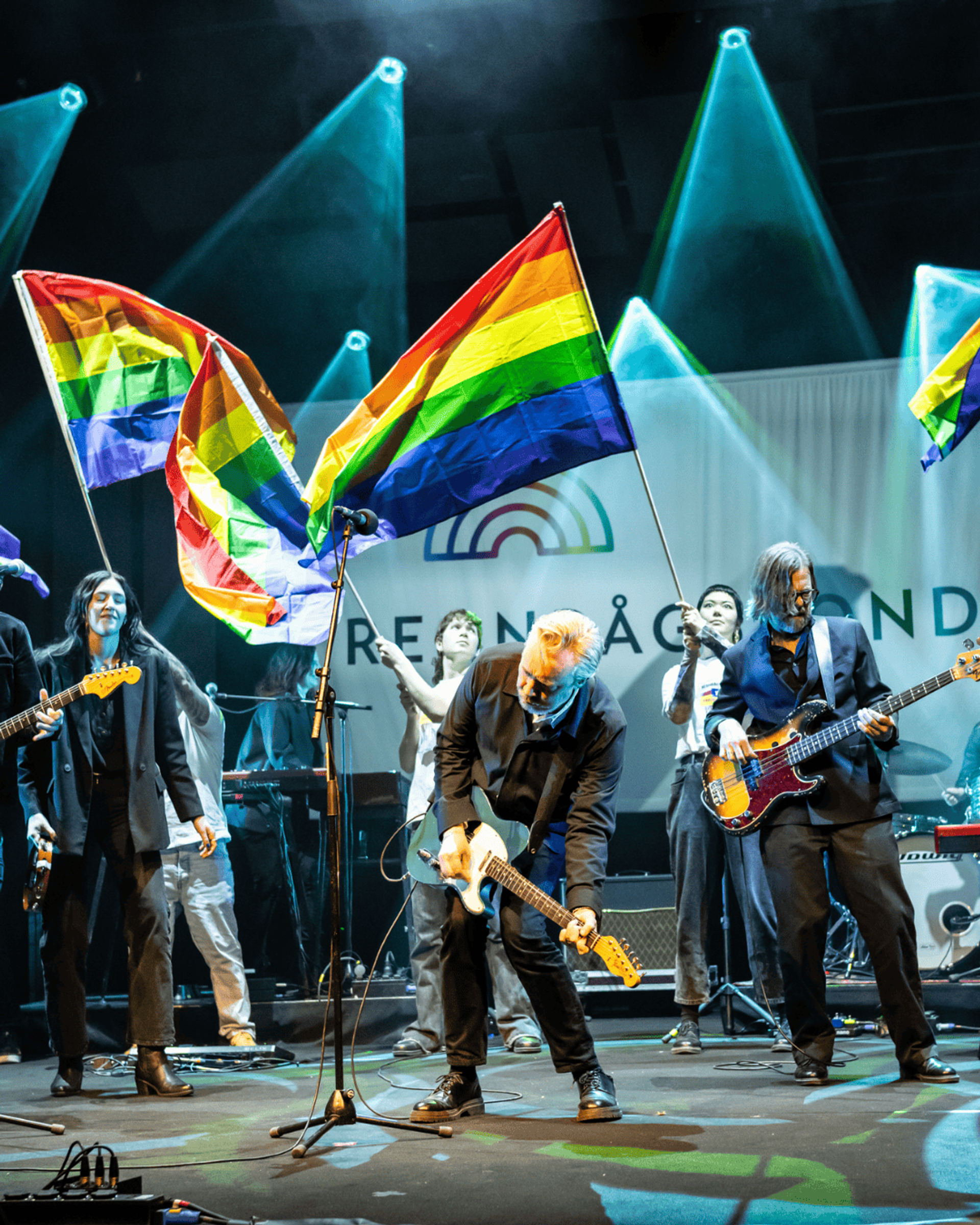 Manér för Regnbågsgalan 2026. Ett band på scen som spelar gitarr och bas med personer som viftar Pride flaggor i bakgrunden.