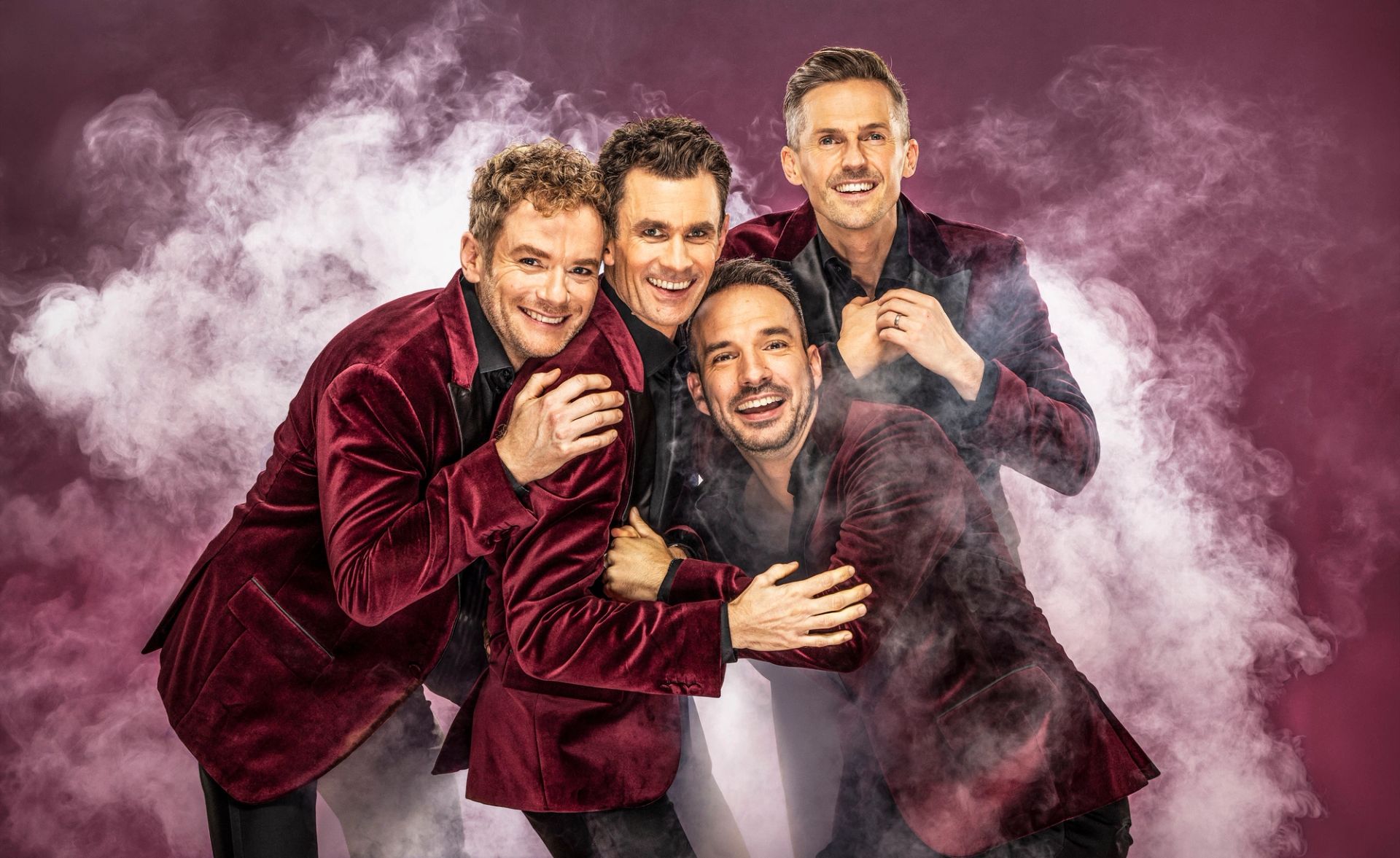 Pressbild för Oh What a Night. Peter Johansson, Bruno Mitsogiannis, David Lindgren och Robert Rydberg har på sig mörkröda sammetskavajer och står mot en mörk bakgrund med vit rök. De tittar in i kameran och ler.