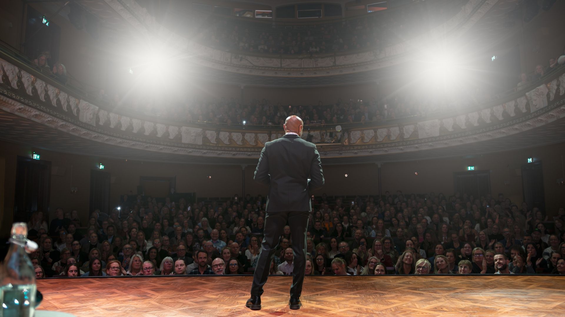 Manér för All Inclusive - Aaron Kroon med gäster. Aaron står på scen med kostym och tittar ut över en publik. Bilden är tagen bakifrån