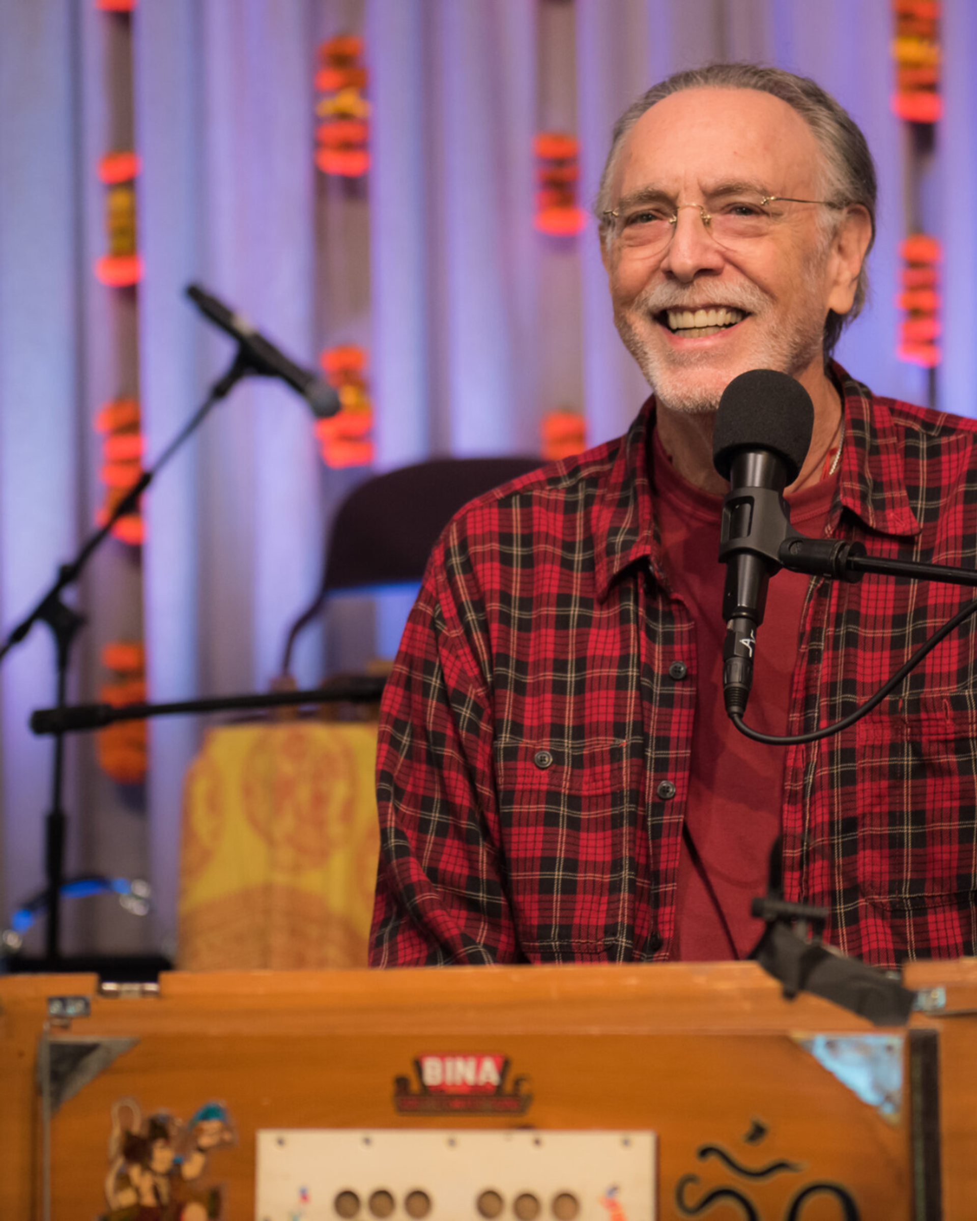Manér bild för föreställningen Kirtan med Krishna Das. Krishna står vid ett podium med en mikrofon framför dig. Bilden är dagen på nära håll. Han har på sig en röd rutig skjorta.