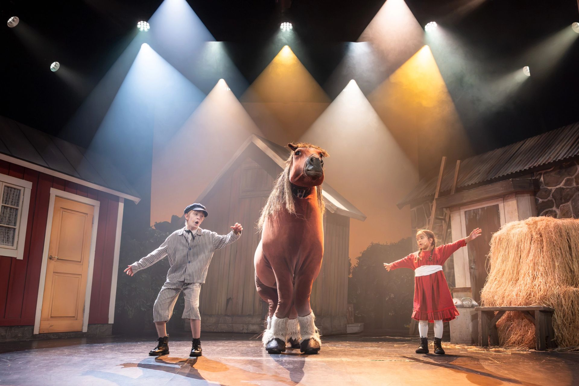 Scenbild från Emil i Lönneberga på Intiman i Stockholm. Emil och Ida står med utsträckta armar på varsin sida om en stor häst, omgivna av gårdshus och höbalar.
