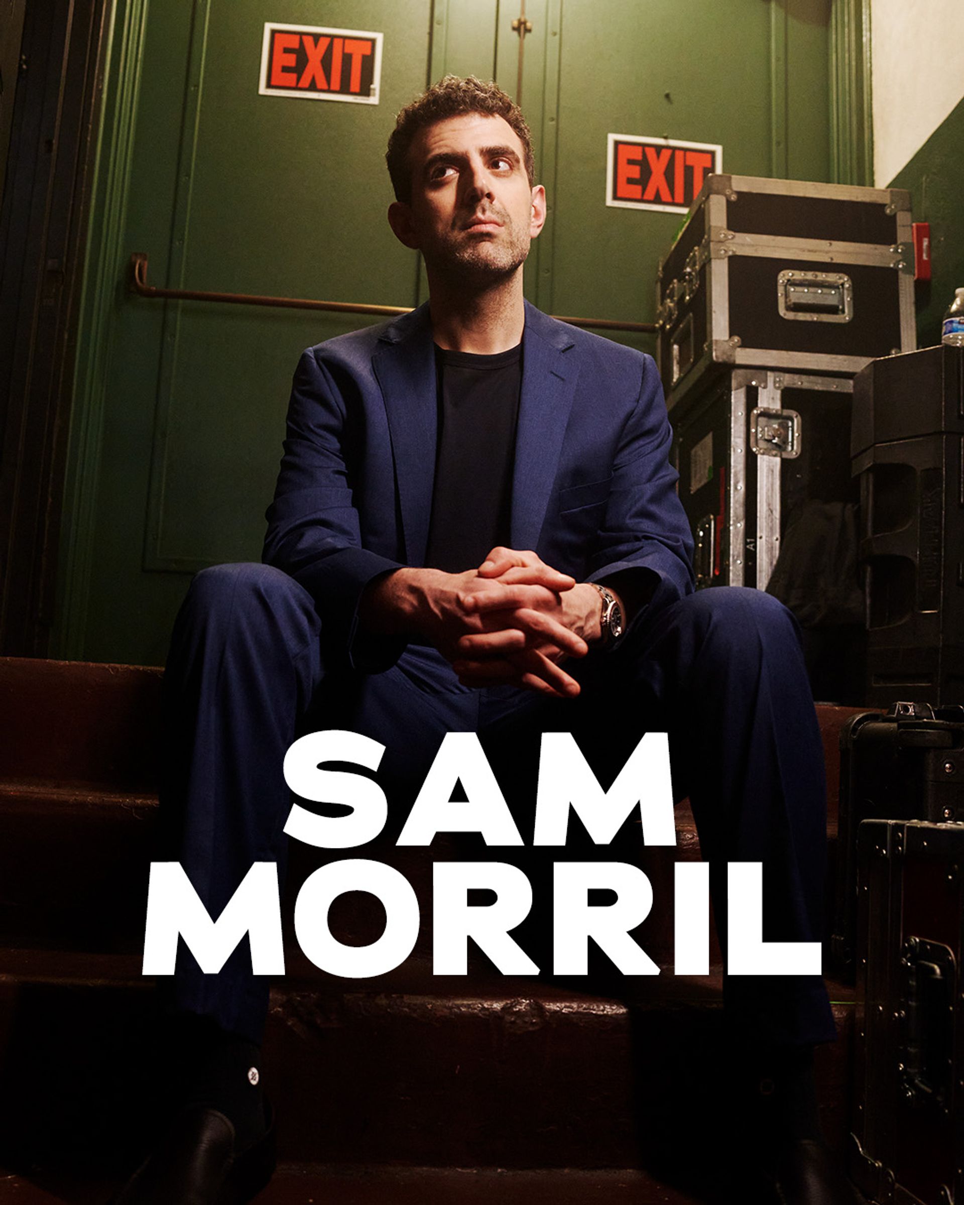 Sam Morril standup show – komiker i blå kostym bakom scen med EXIT-skyltar, porträtt inför liveföreställning