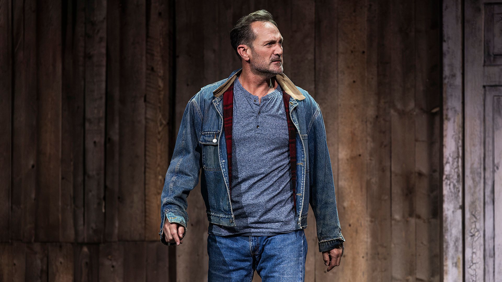 Scenbild från musikalen Så som i himmelen på Lorensbergsteatern i Göteborg. Robin Stegmar står i jeans, blå tröja och jeansjacka framför en rustik trävägg och ser allvarligt åt sidan.