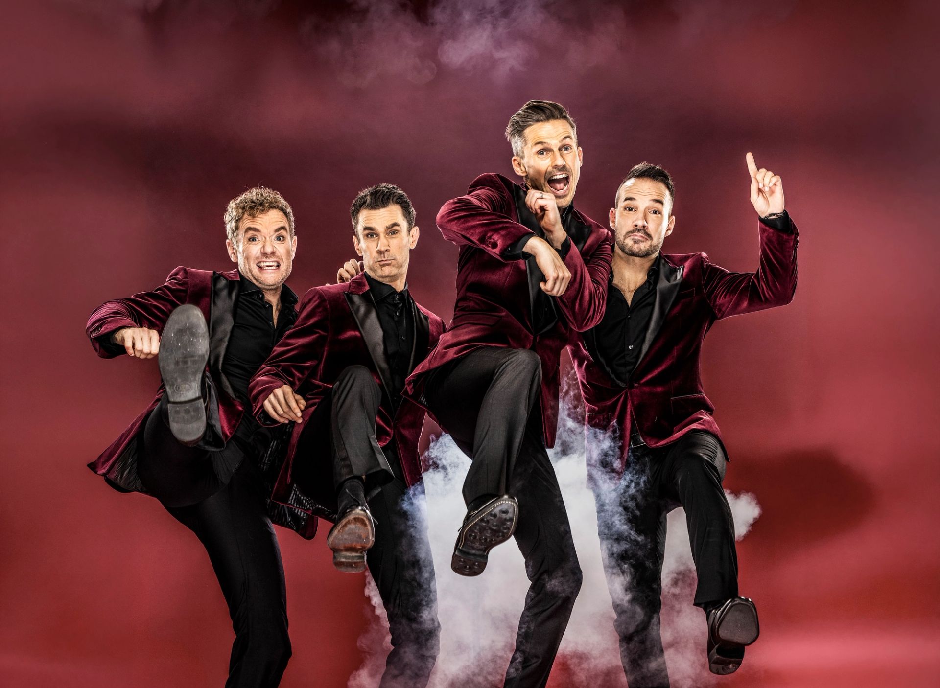 Pressbild för Oh What a Night. Peter Johansson, Bruno Mitsogiannis, David Lindgren och Robert Rydberg har på sig mörkröda sammetskavajer och står mot en mörk bakgrund med vit rök. Alla fyra hoppas och gör en spark mot kameran.