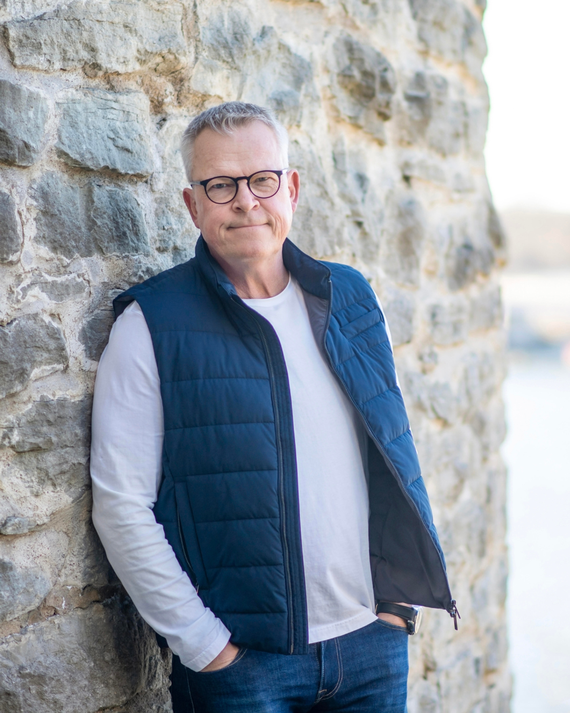 Janne Andersson poserar framför en stenvägg. Han bär ljus långärmad tröja och blå dun väst.