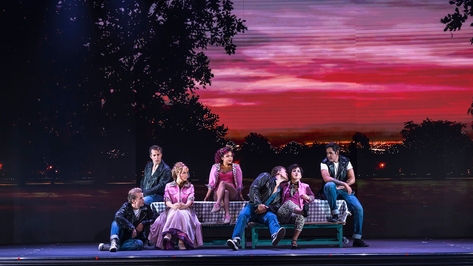 T-Birds och Pink Ladies i en scen ur Grease The Musical på Rondo i Göteborg. Grupperna sitter tillsammans i färgstarka 50-talskläder framför en dramatisk solnedgång.