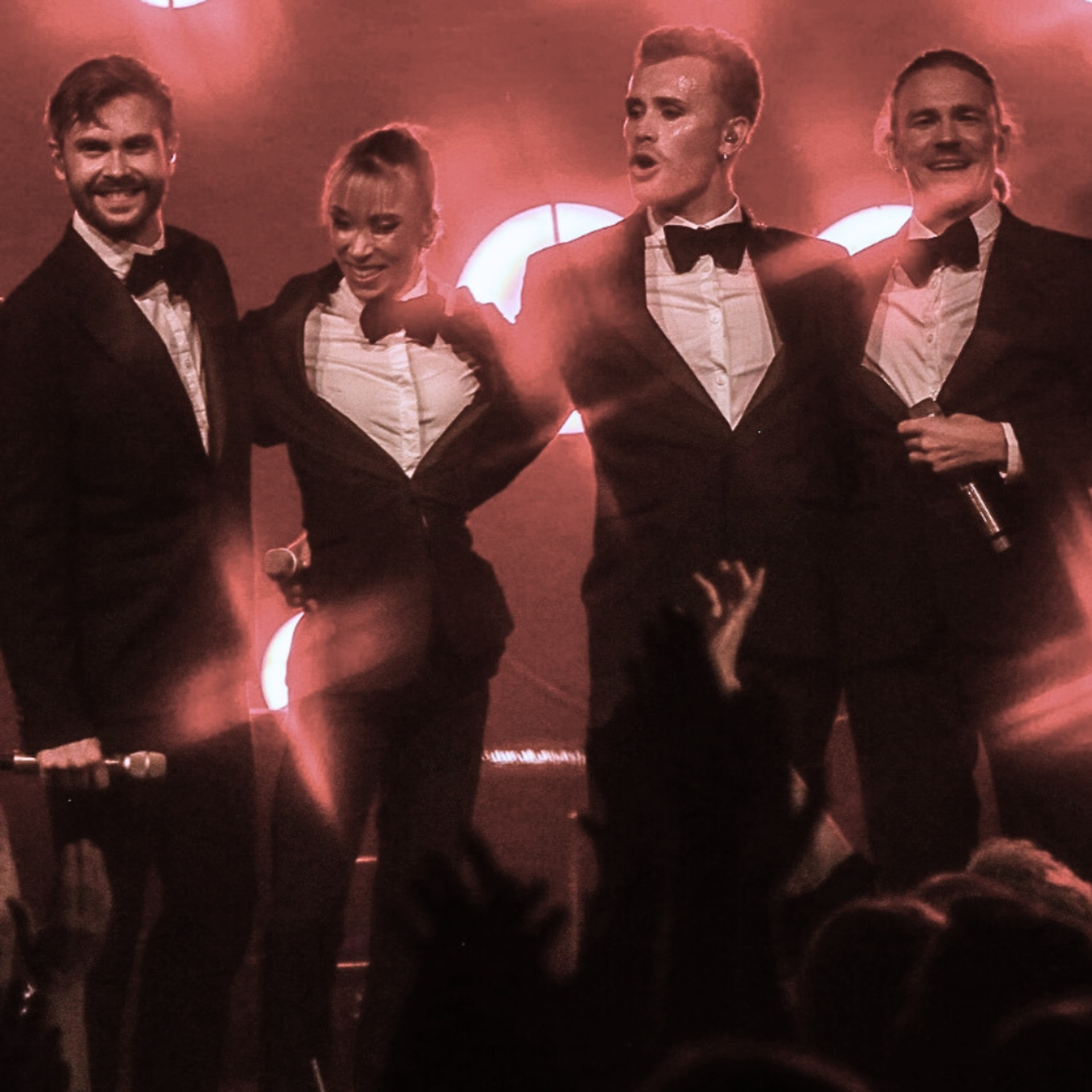 Wallmans show. Fyra artister i kostym som står på scen och ler ut emot publiken. Ljuset är rött.