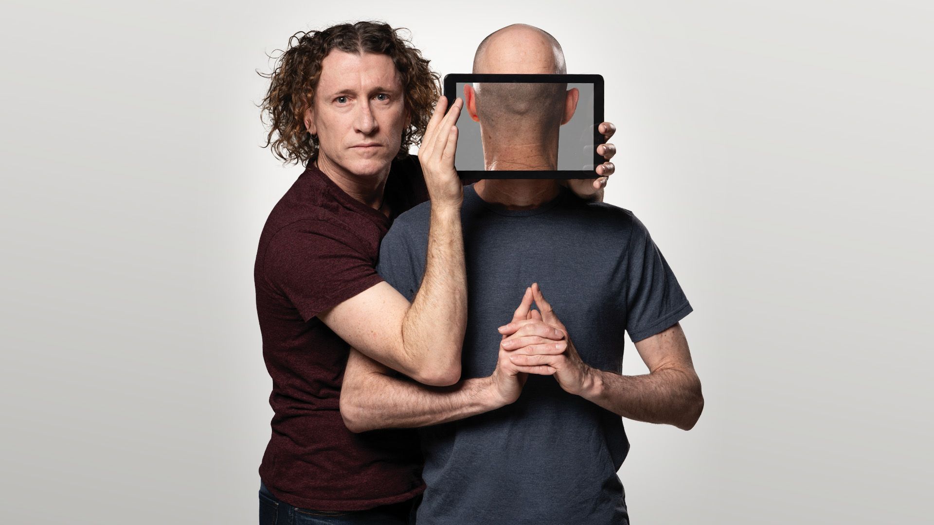 Manér bild för The Umbilical Brothers - The Distraction. The Umbilical Brothers står båda med kroppen mot kameran men den ena brodern håller en iPad framför den andras ansikte så det ser ut som att han står med ryggen mor kameran.