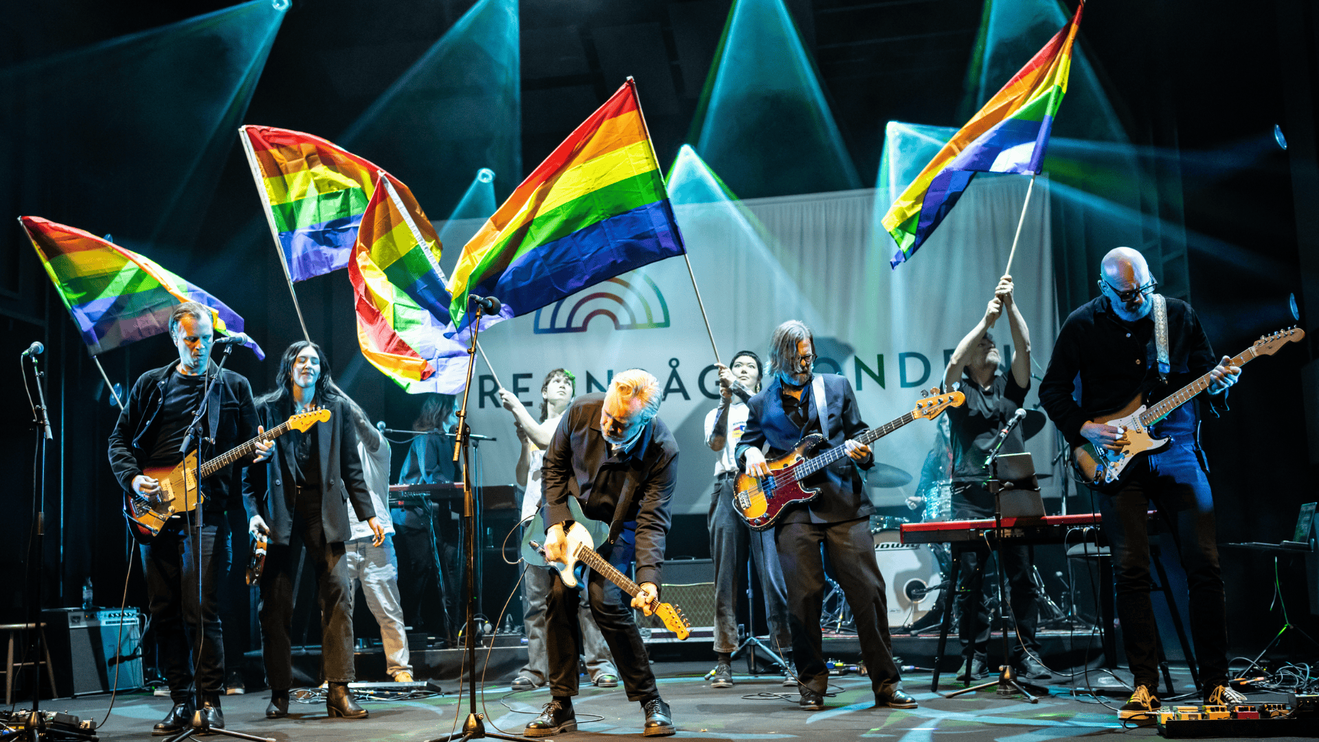 Manér för Regnbågsgalan 2026. Ett band på scen som spelar gitarr och bas med personer som viftar Pride flaggor i bakgrunden.