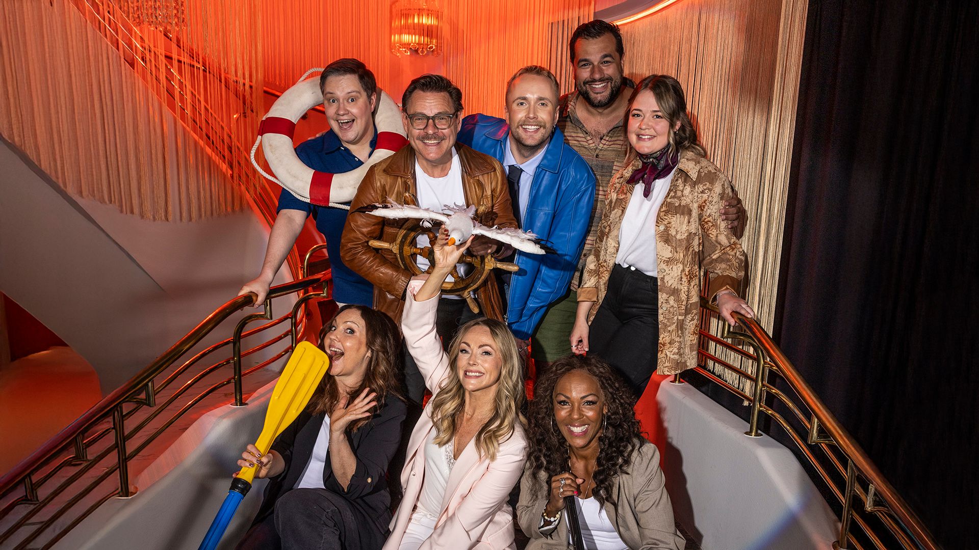 Pressbild med Linda Ohlsson, Edvin Törnblom, Edward af Sillén, Alma Bengtsson, Anton Lundqvist, Martin Redhe Nord, Kim Sulocki, Hanna Hedlund och Kayo från föreställningen Titanique på Börsen i Stockholm. Ensemblen poserar tillsammans i en trappa med maritim rekvisita som livboj, båtratt, mås och paddel, i en varm orangebelyst miljö.