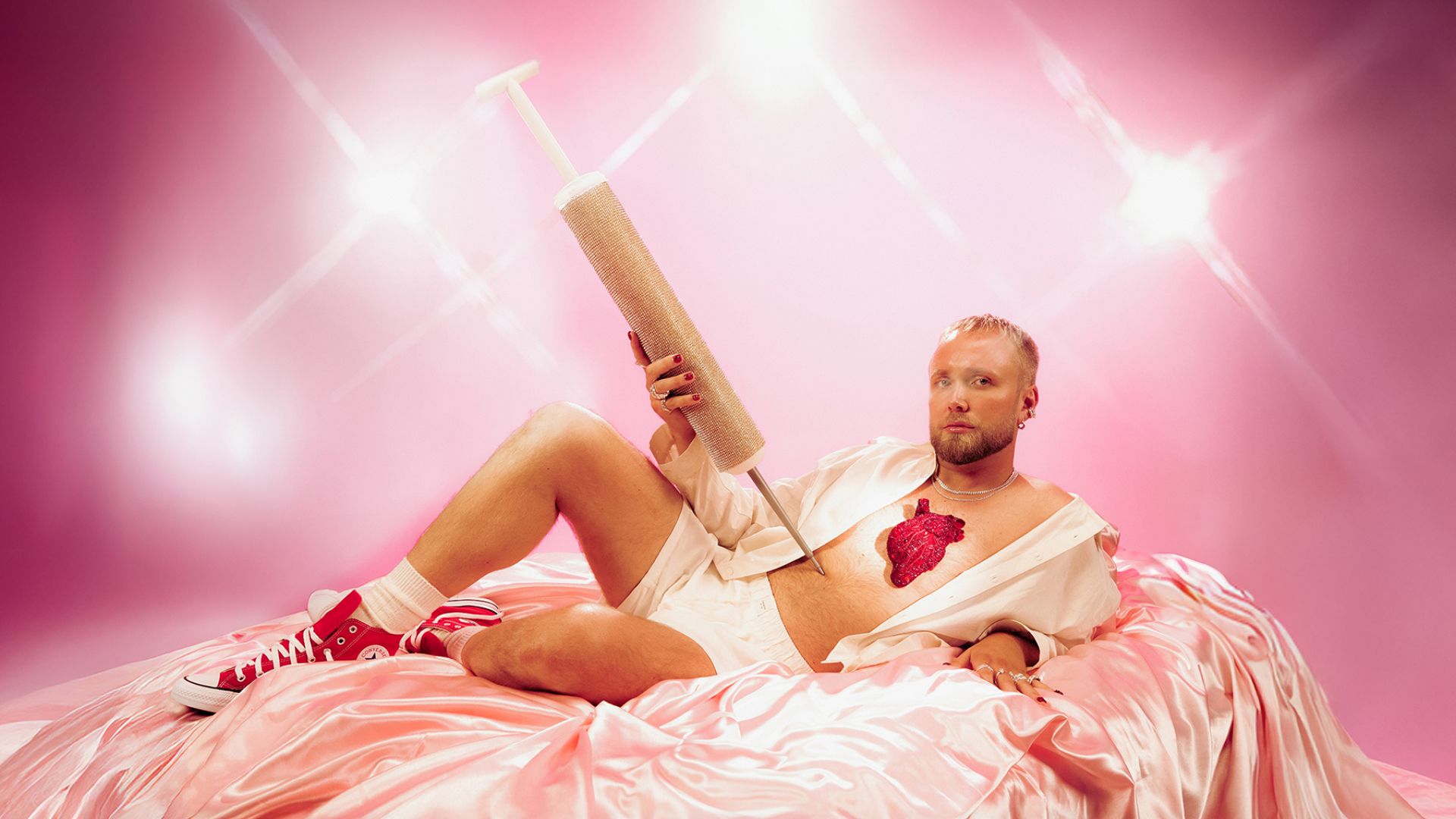 Manér bild för Eviga Edvin med Edvin Törnblom. Han ligger på en säng med siden lakan iklädd siden pyjamas shorts och en skjorta. Hela bilden går i rosa färger.