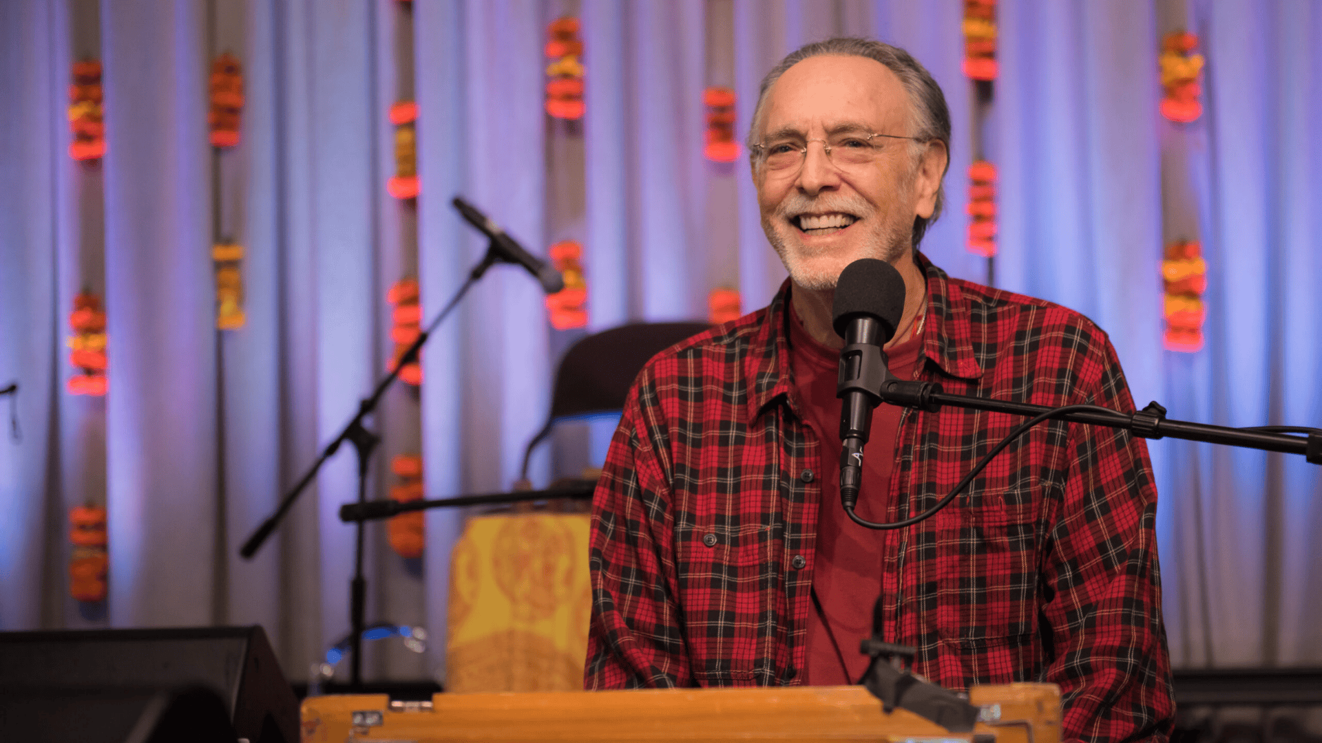 Manér bild för föreställningen Kirtan med Krishna Das. Krishna står vid ett podium med en mikrofon framför dig. Bilden är dagen på nära håll. Han har på sig en röd rutig skjorta.