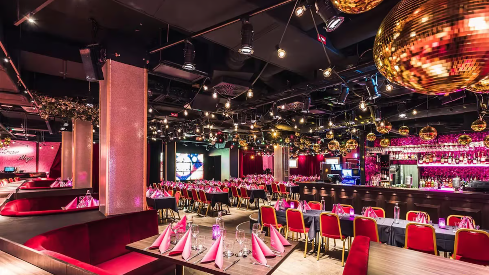 Interiör av Golden Hits i Stockholm med långbord dukade för middag, röda stolar, rosa belysning, stora discokulor i taket och en bar i bakgrunden.