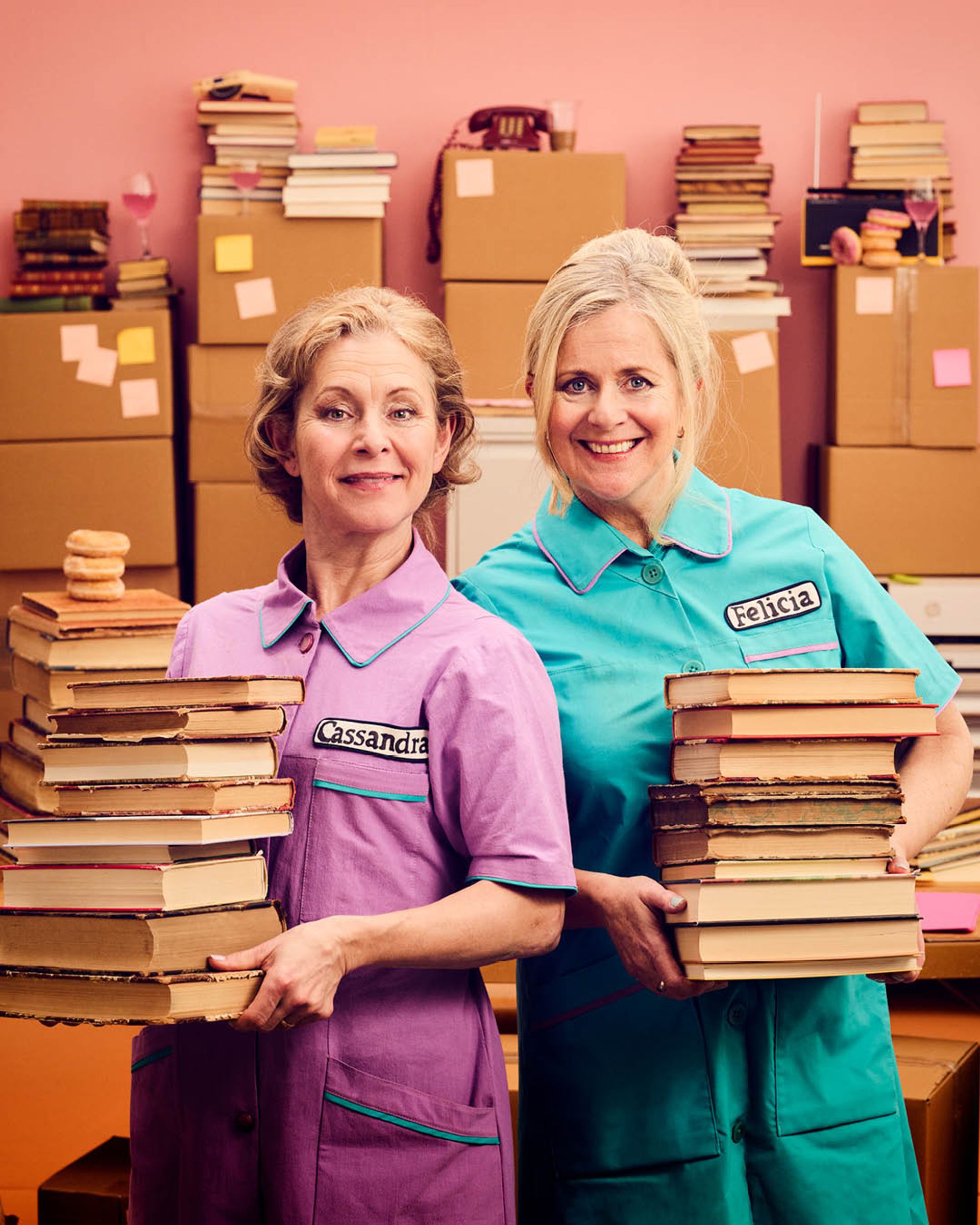 Affisch för musikalen Nån måste göra det med Gunilla Backman och Helen Sjöholm, som spelas på Intiman i Stockholm 7–14 april 2027. Kvinnor i turkosa och lila arbetskläder står bland kartonger, böcker och kontorsmaterial mot en rosa bakgrund.