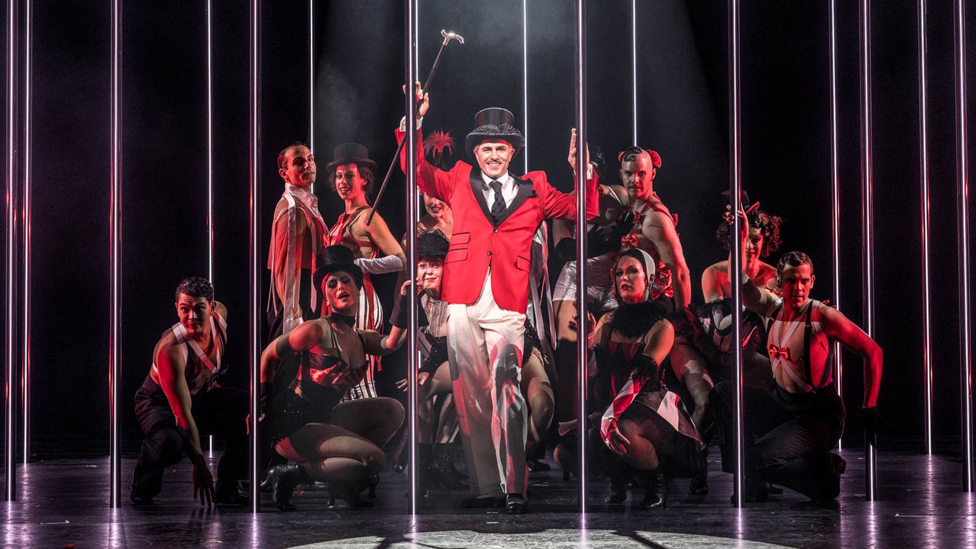 Scenbild från musikalen Chicago på Oscarsteatern i Stockholm. Peter Jöback står i mitten av ensemblen, klädd i röd kavaj, vit kostym och svart hatt, med en käpp höjd i ena handen. Runt honom poserar dansare i svartvita scenkostymer bland höga vertikala ljusstavar, med dramatisk röd och vit scenbelysning mot en mörk bakgrund. Chicago spelas på Oscarsteatern 16 september–19 december.
