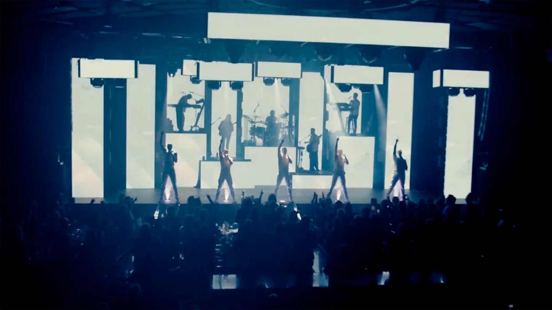 Bild tagen under ett framträdande på showen A Show Larger Than Life. 5 av artisterna står på scen med sin vänstra arm rakt upp i luften.