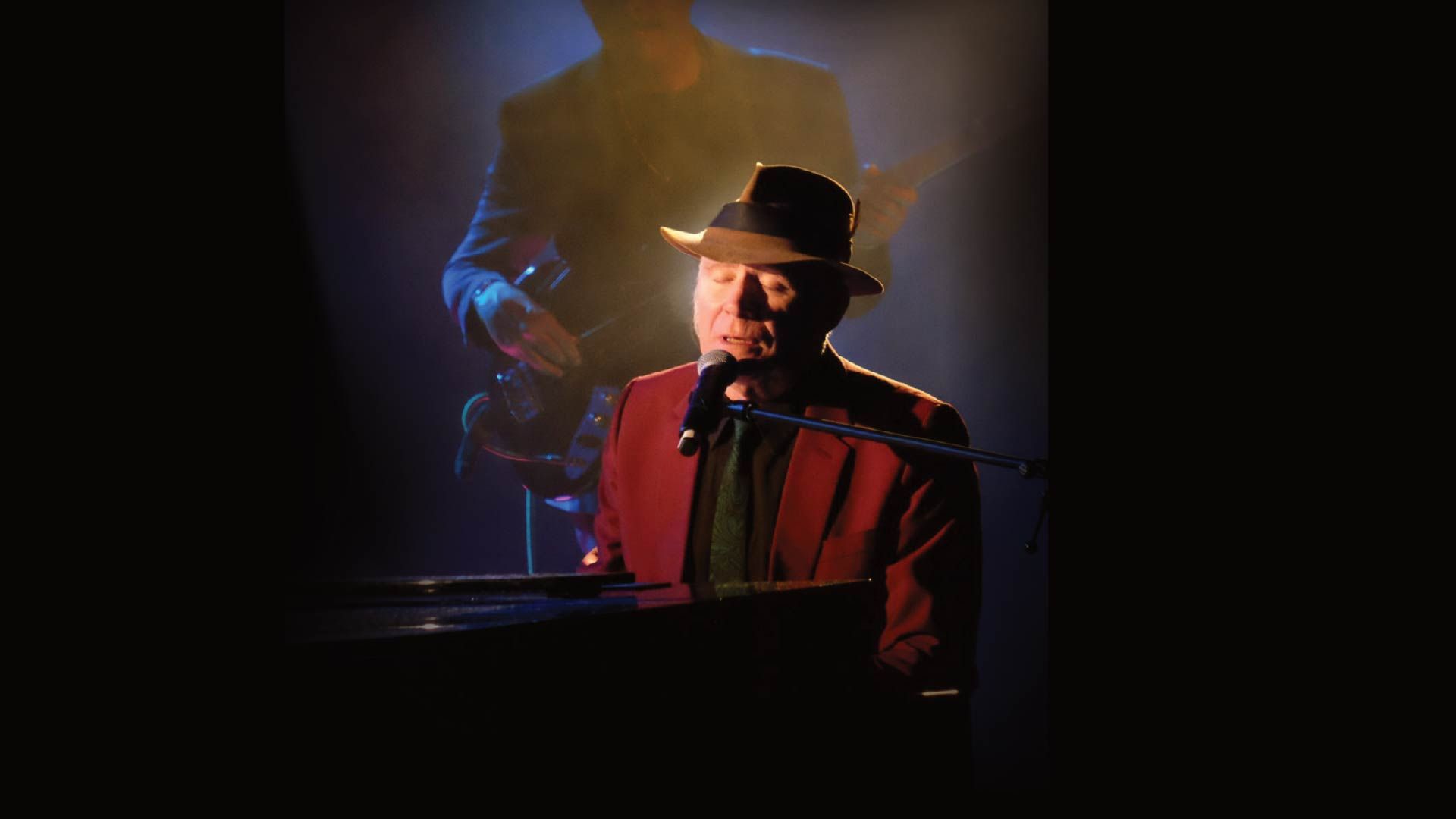 Manér bild för konserten My Leonard Cohen som föreställer Stewart D'Arrietta när han sitter och sjunger vid ett piano under en strålkastare.