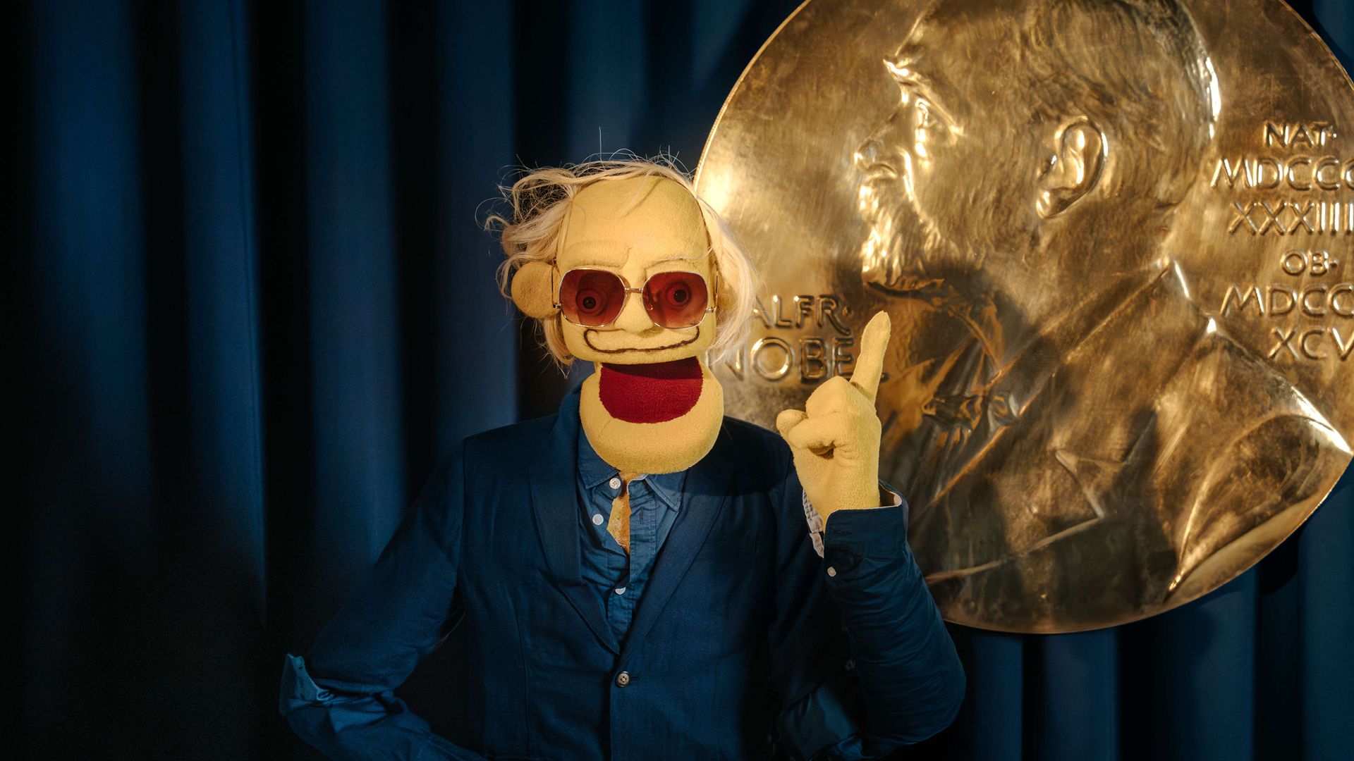 Manérbild för Crash Course i årets Nobelpriser – bild på en karaktär i blå kostym framför en bakgrund med Nobelpriset. Evenemanget presenteras av Nobel Prize Museum och kombinerar humor och kunskap inför Nobelpriset 2025.