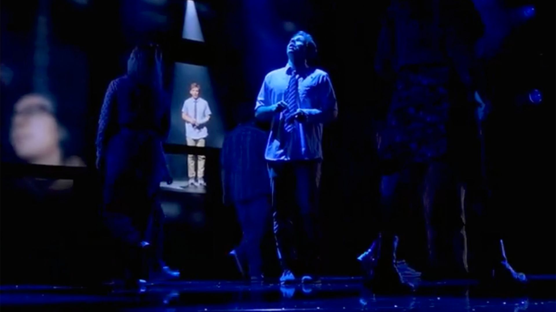Bild från föreställning Dear Evan Hansen. Skådespelaren som spelar Evan står i mitten i bilden och tittar upp i luften. Bakom honom lyser det upp en helkropps bild som ska gestalta Evan som yngre.