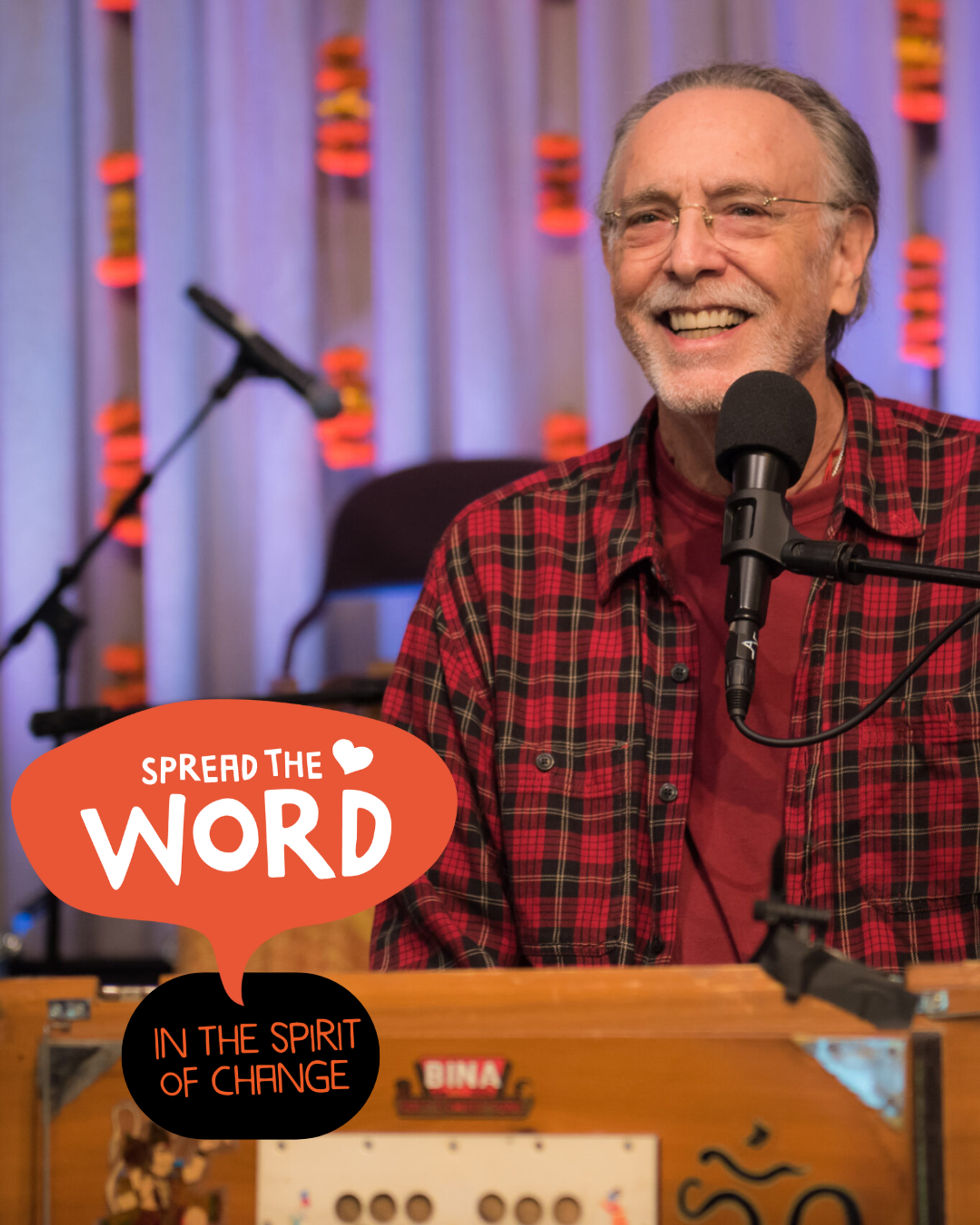 Manér bild för föreställningen Kirtan med Krishna Das. Krishna står vid ett podium med en mikrofon framför dig. Bilden är dagen på nära håll. Han har på sig en röd rutig skjorta.