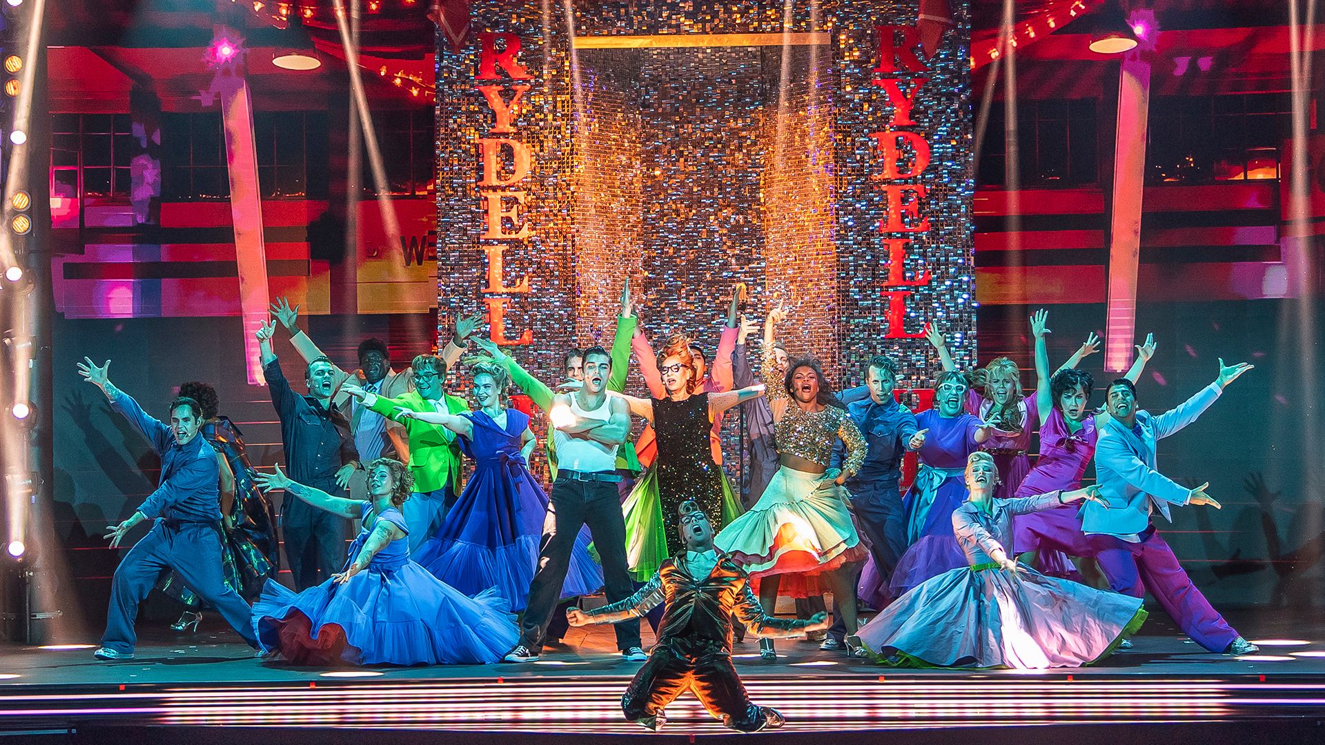 Ensemble från Grease The Musical på Rondo i Göteborg i ett färgsprakande shownummer. Ensemblen dansar i färgstarka scenkläder framför en glittrande scenografi med texten ”Rydell”.